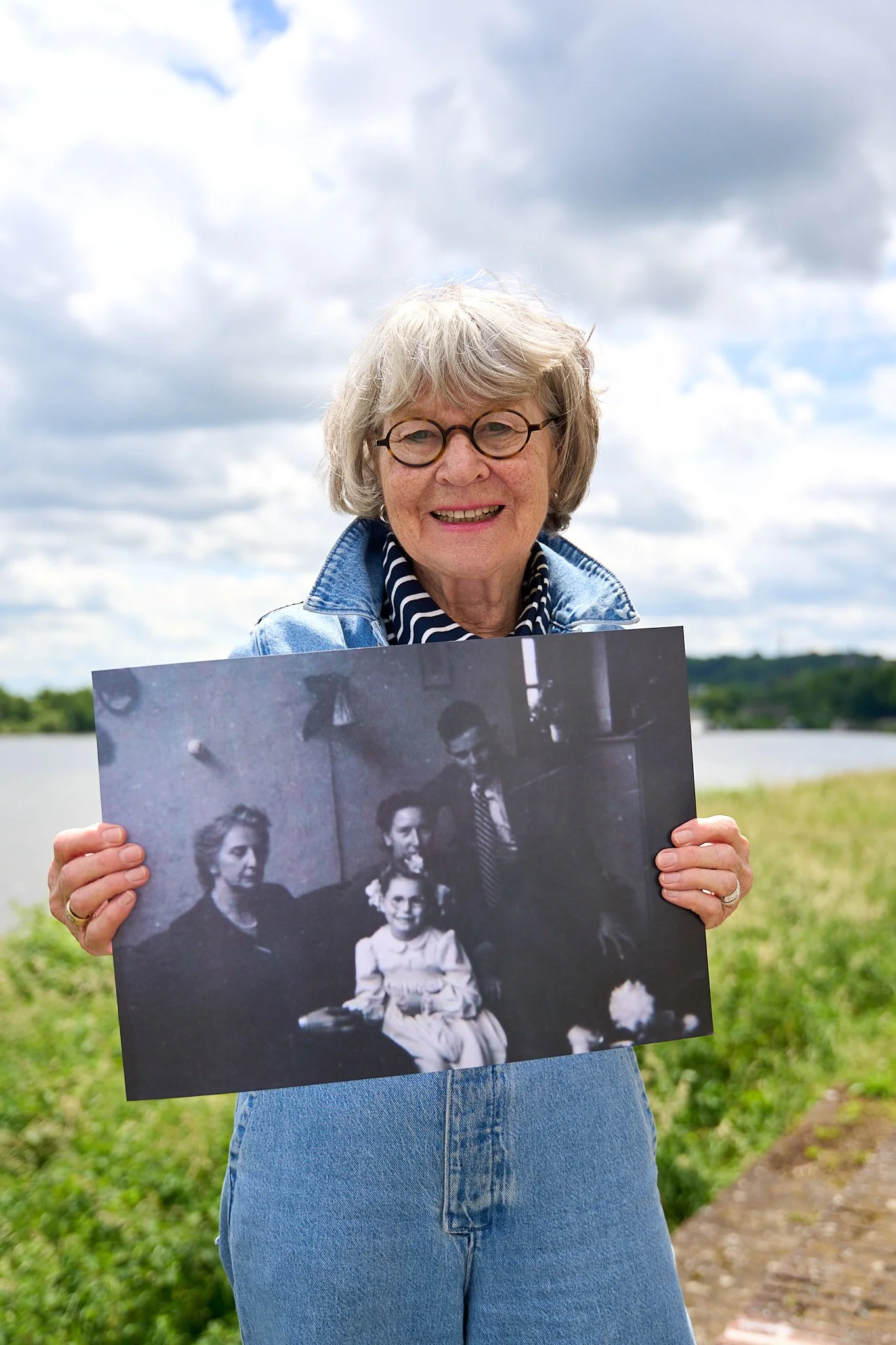 A smiling woman with glasses and a striped shirt holding a black-and-white family photograph outdoors near a lake with cloudy sky.