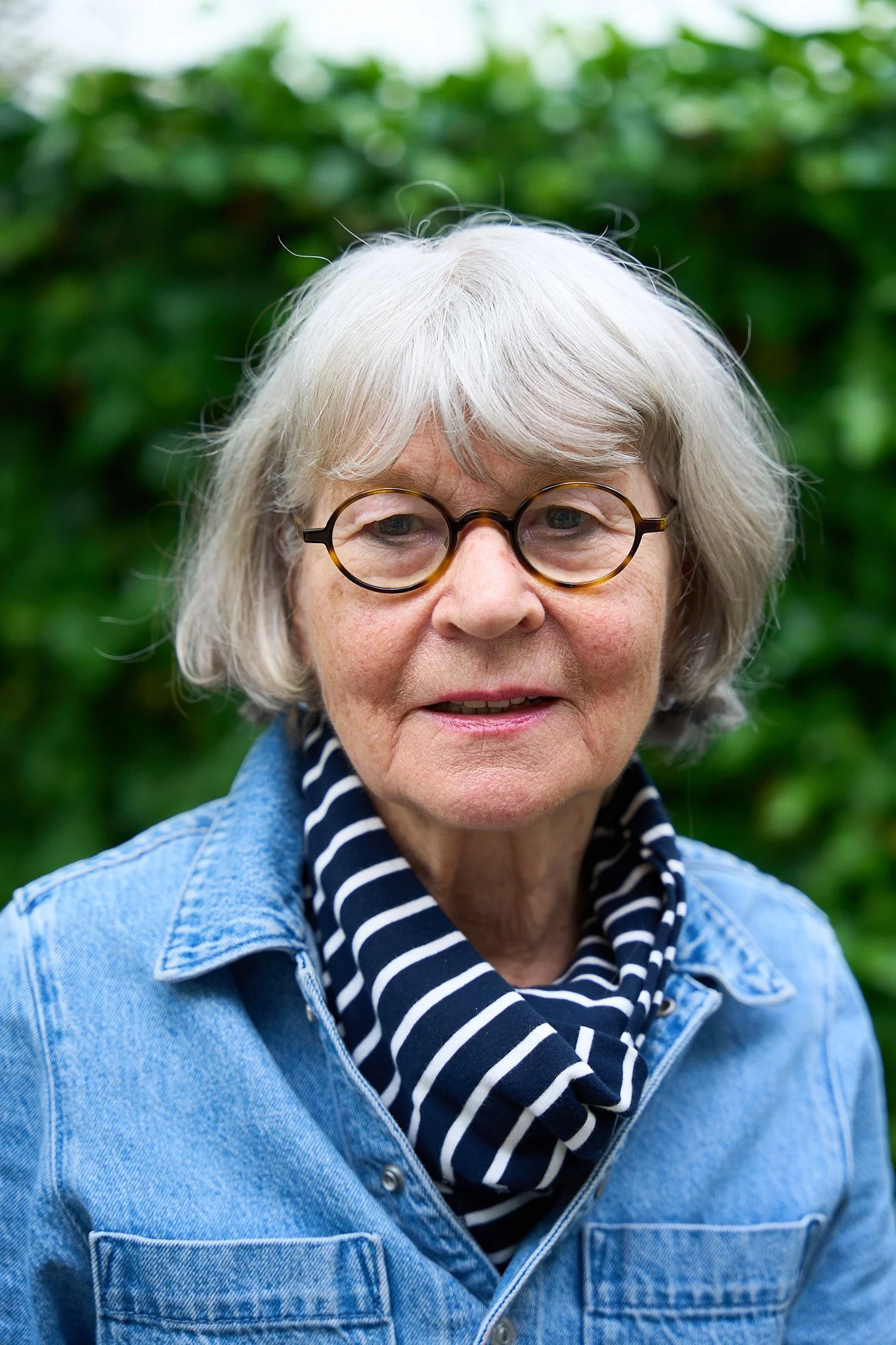 An elderly woman with gray hair and glasses, wearing a denim jacket and a black-and-white striped scarf, standing outdoors with green foliage in the background.