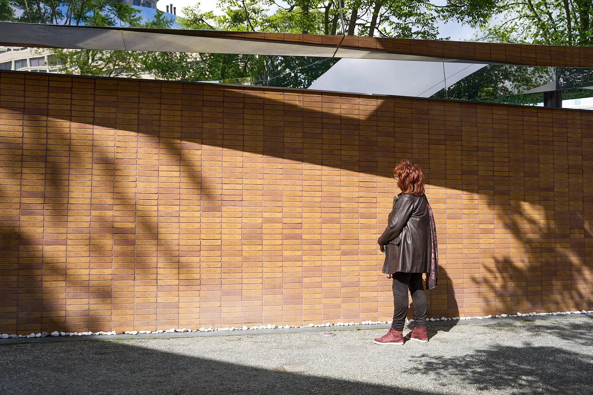 A woman with curly red hair wearing a black leather jacket, black pants, and red boots standing in front of a brick wall with shadows of other trees cast on it, in an outdoor area during daytime.