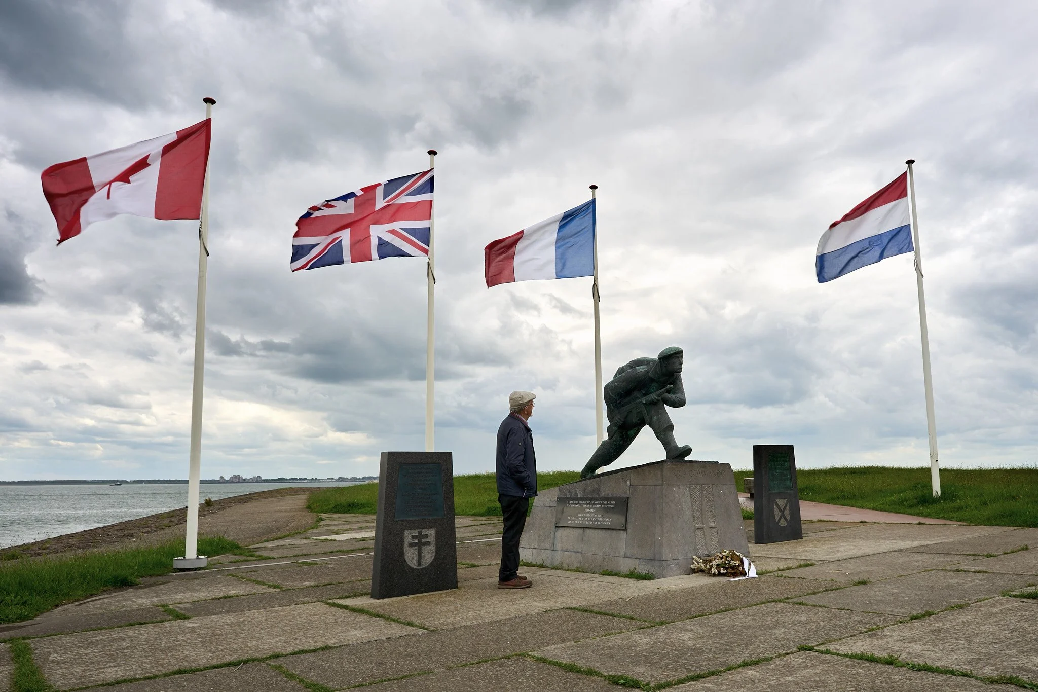 A man standing in front of a war memorial with a statue of a soldier, surrounded by four flags of Canada, the United Kingdom, France, and the Netherlands, on a cloudy day by a body of water.