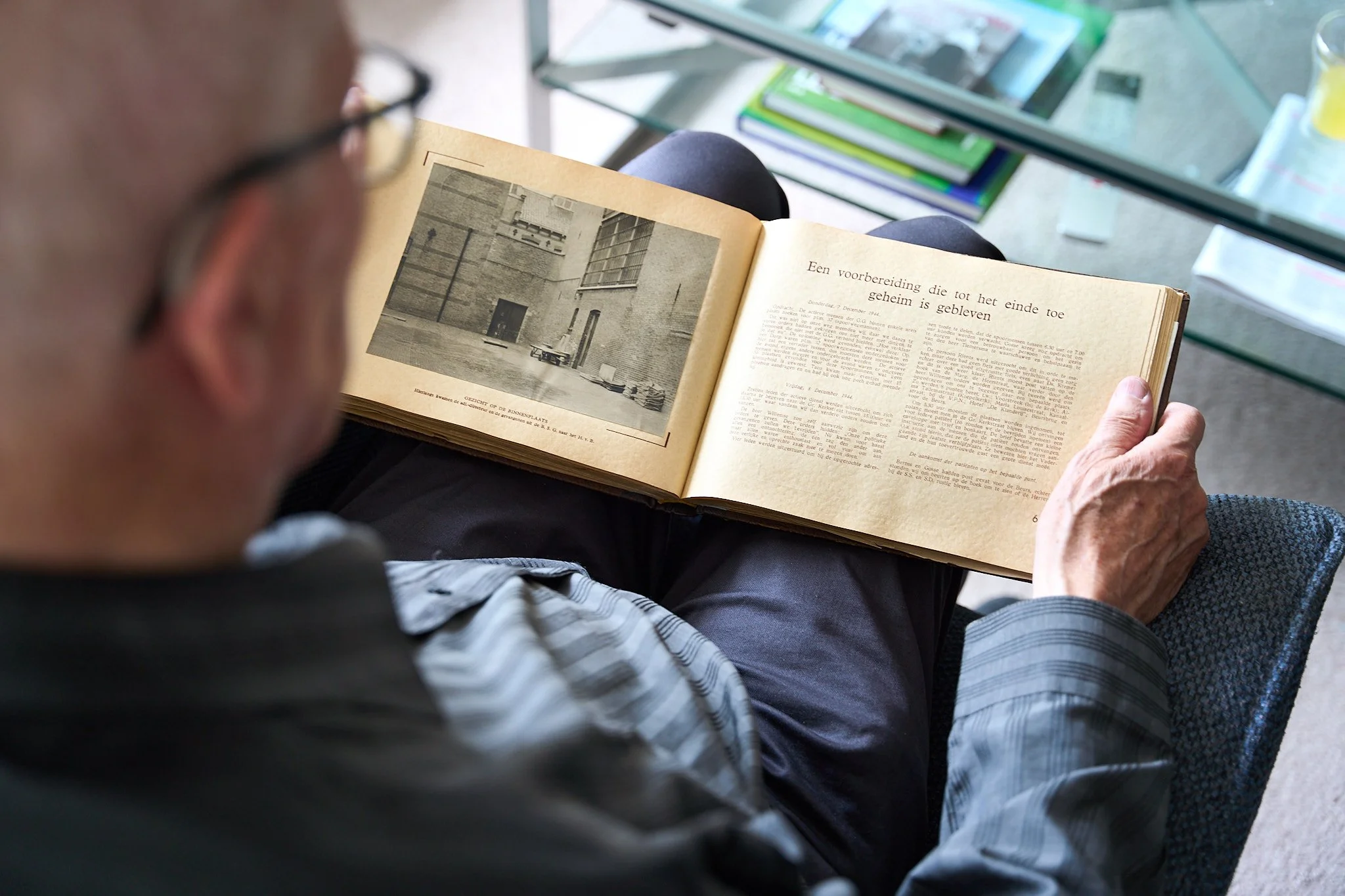 An elderly man with glasses reading a large, open, vintage photo album on his lap, surrounded by magazines and a glass table with books in a bright room.