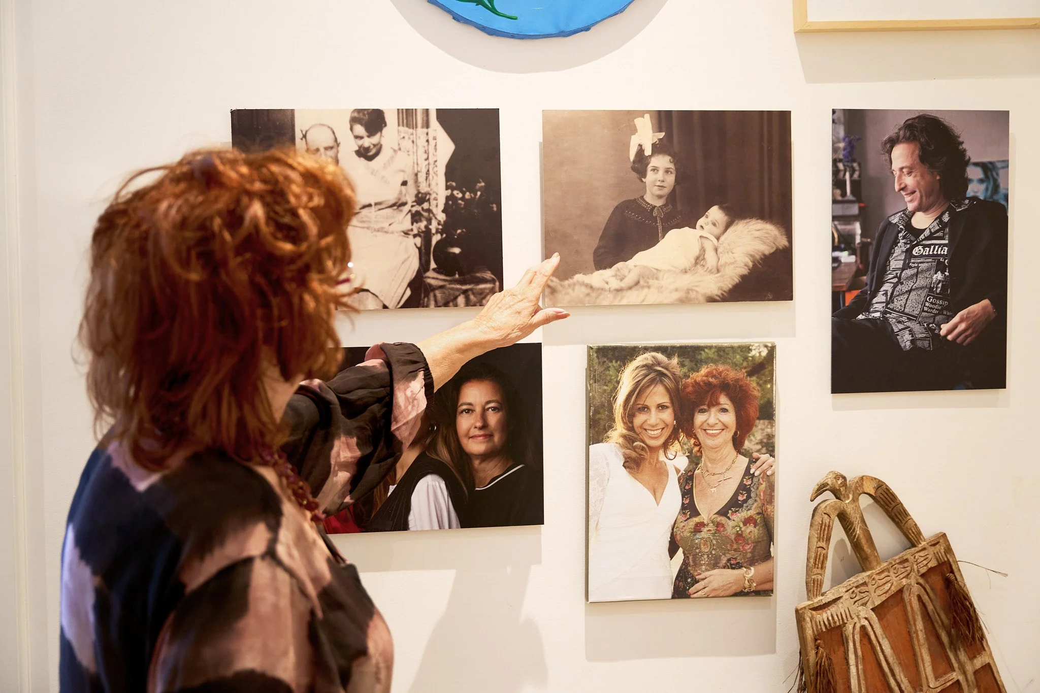 A woman with red hair points at a photograph on a white wall with other photos. The photos include black-and-white and color images of people, including a woman with a child, and two women smiling outdoors. There is a carved wooden item leaning again