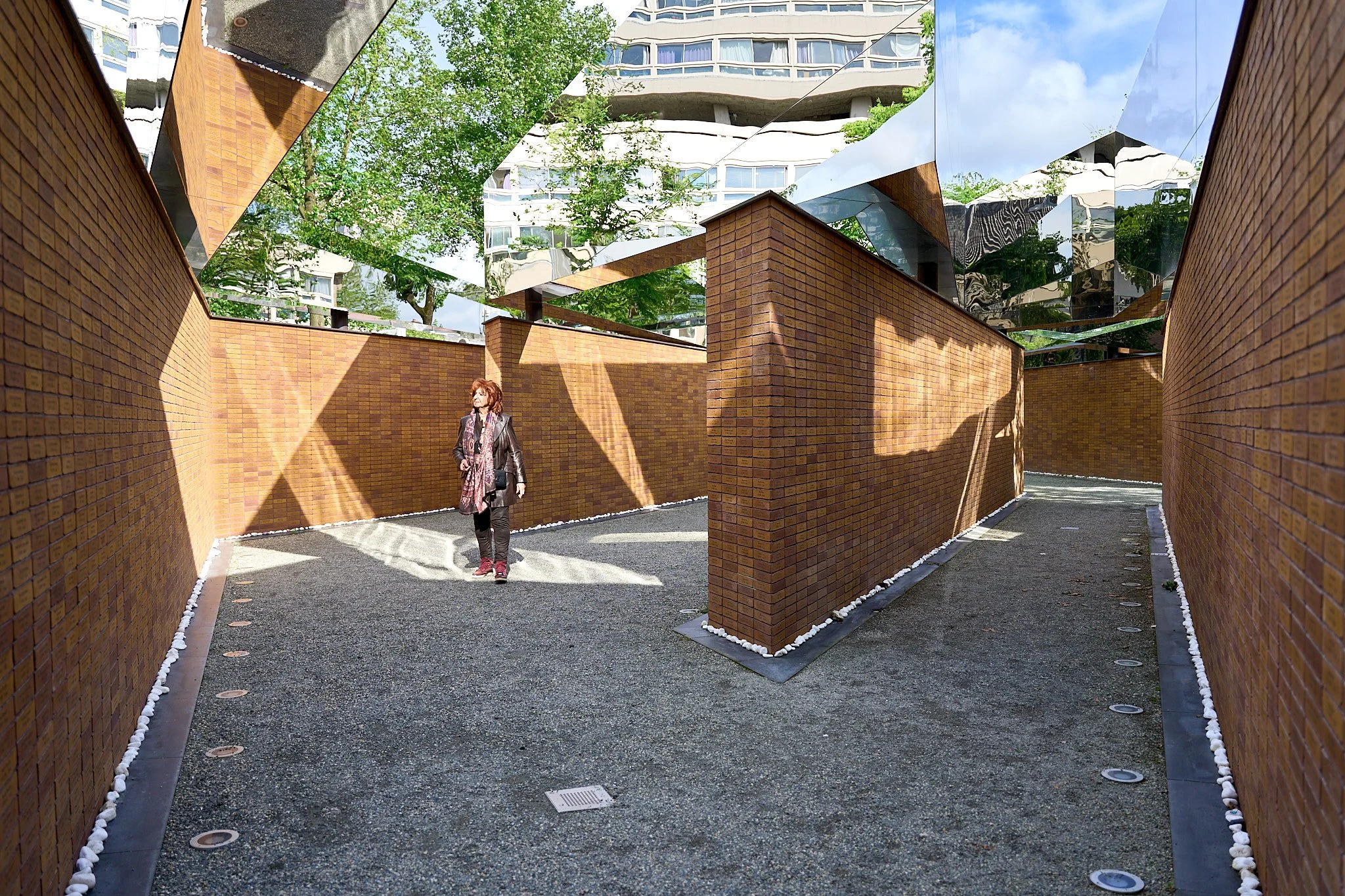 A woman walking through a modern architectural outdoor space with high brick walls, reflective metal panels on the ceiling, and trees in the background, with shadows cast on the ground.