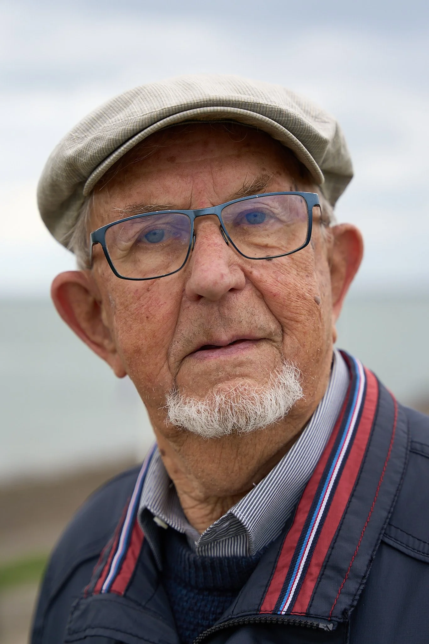 Close-up of an elderly man with glasses and a beard, wearing a cap, outdoor setting with cloudy sky background.