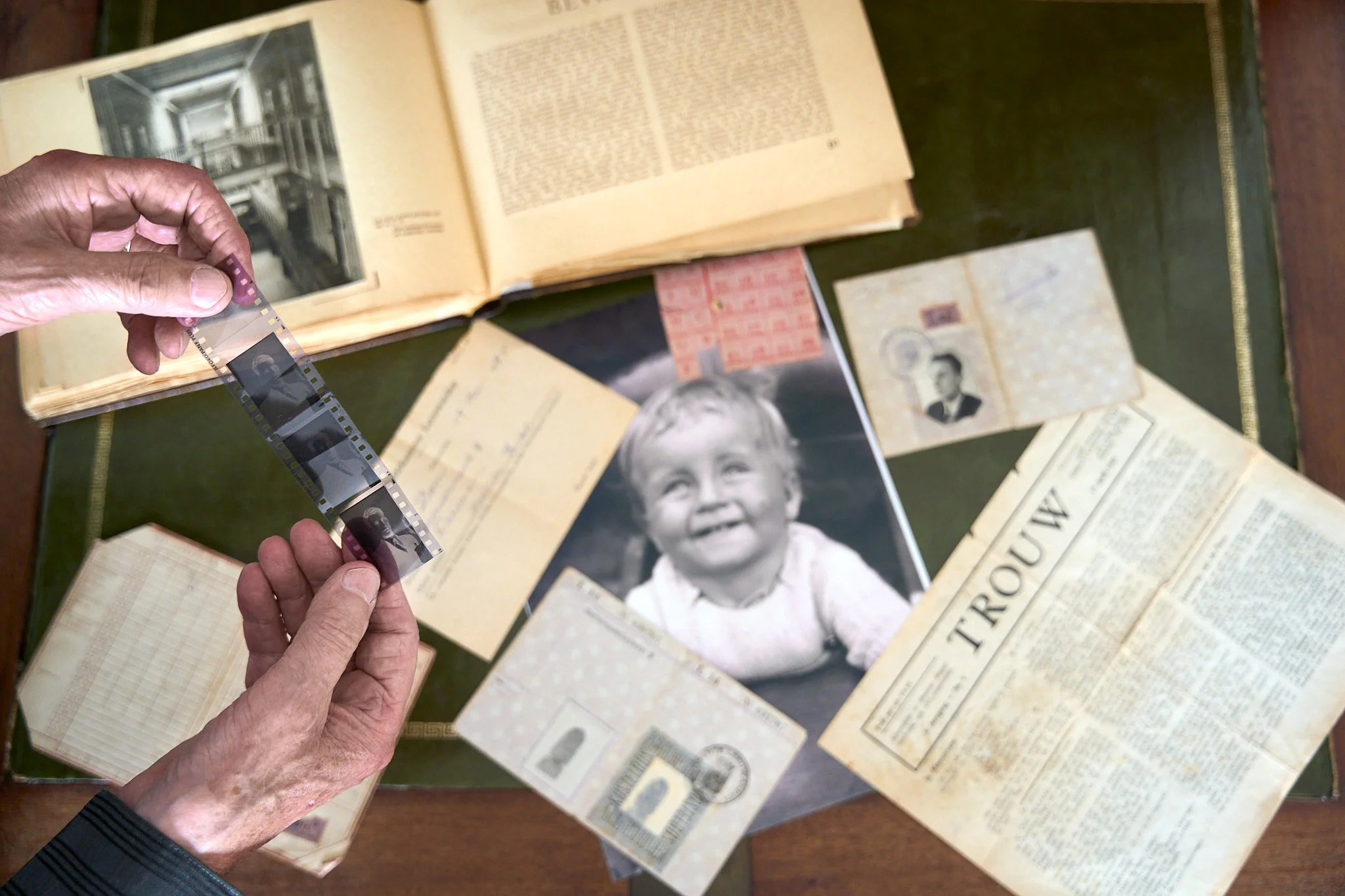 A person holds a strip of black-and-white photographic negatives over a table with vintage photographs, documents, and a newspaper, including a smiling child in a black-and-white photograph.