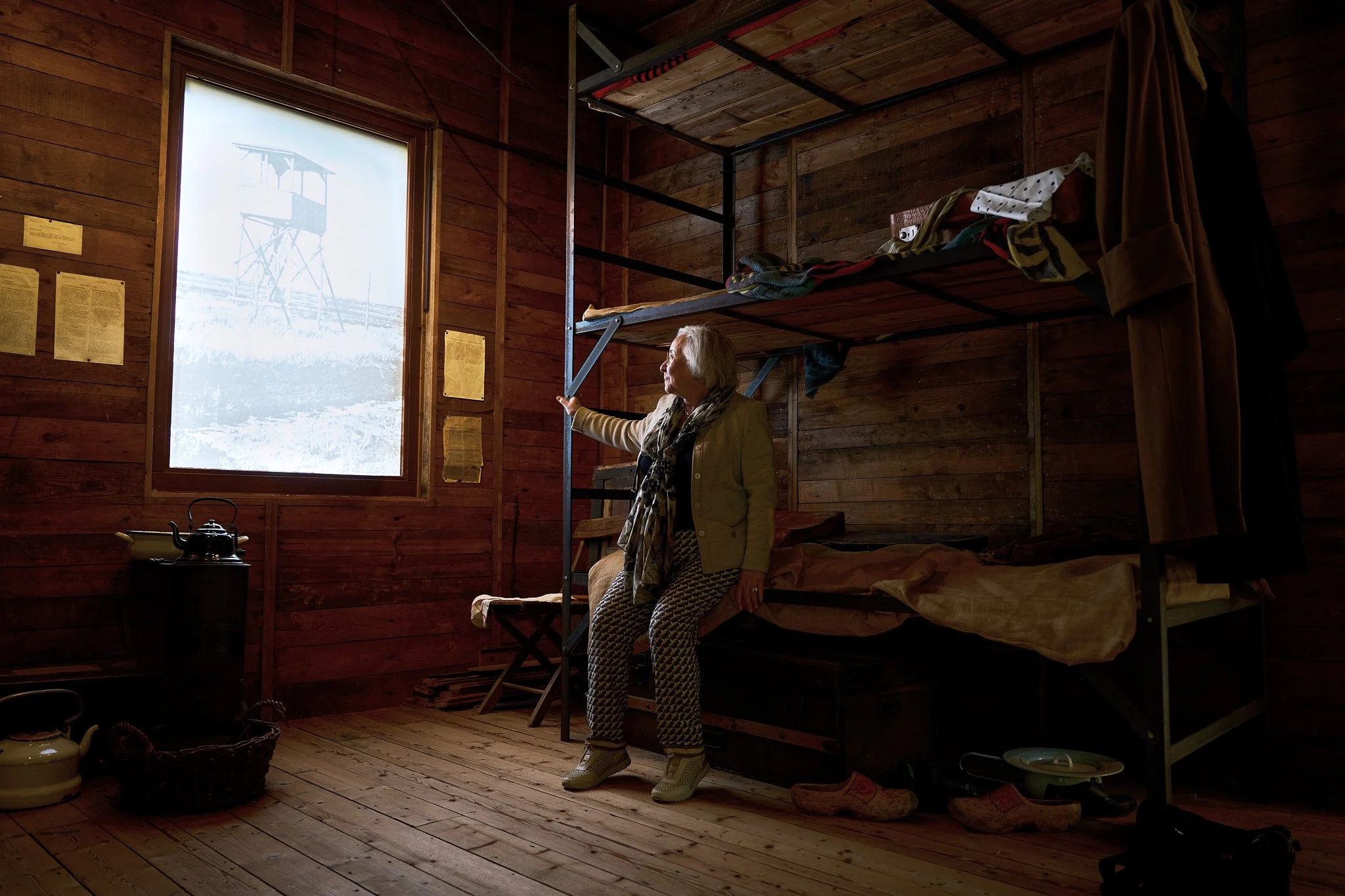 An elderly woman with gray hair sitting on a bed inside a wooden cabin, pointing at a watchtower outside the window, with a warm natural light coming through.