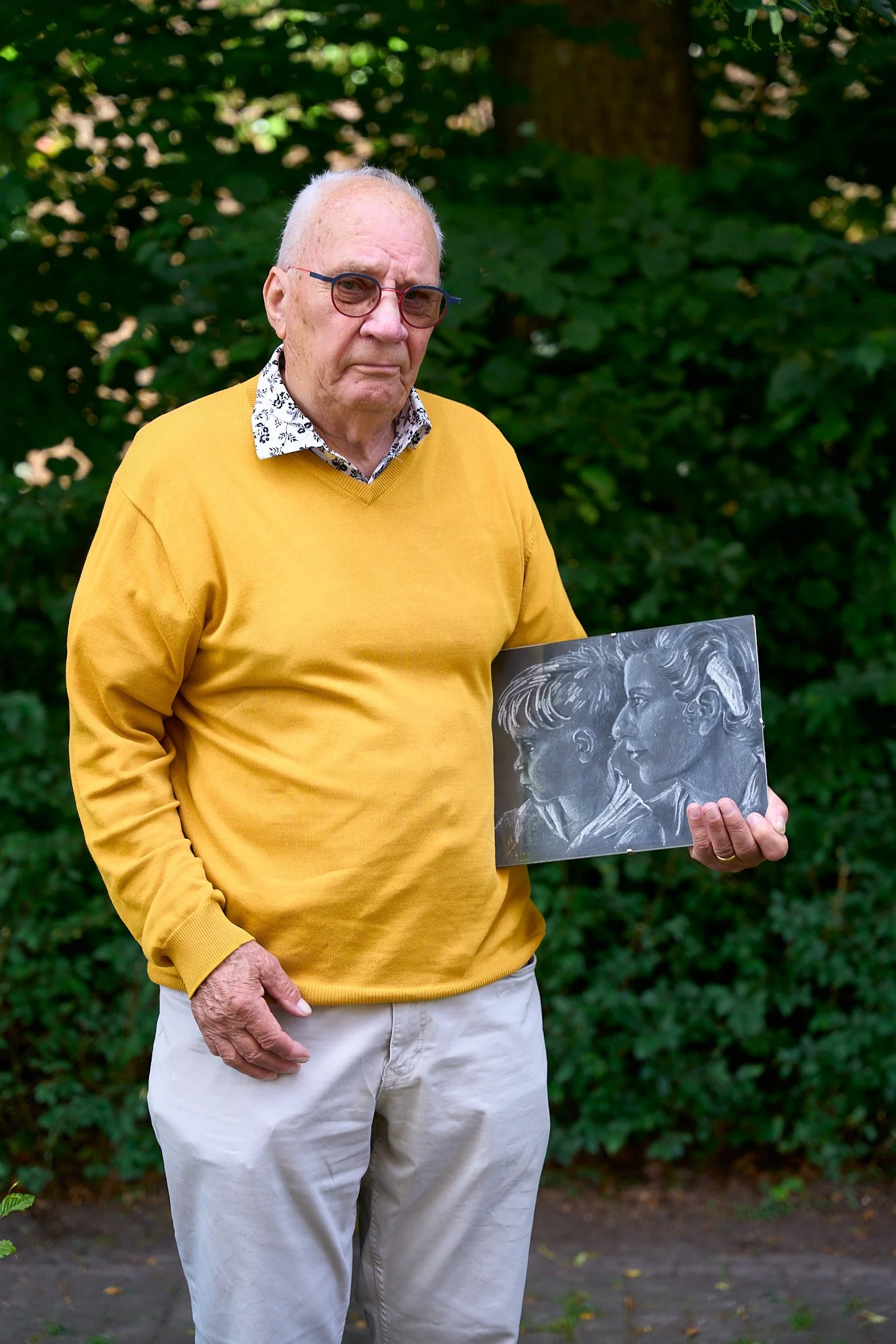 An elderly man with glasses wearing a yellow sweater and white pants stands outdoors holding a piece of artwork that depicts two children in profile, rendered in white chalk on a black background.