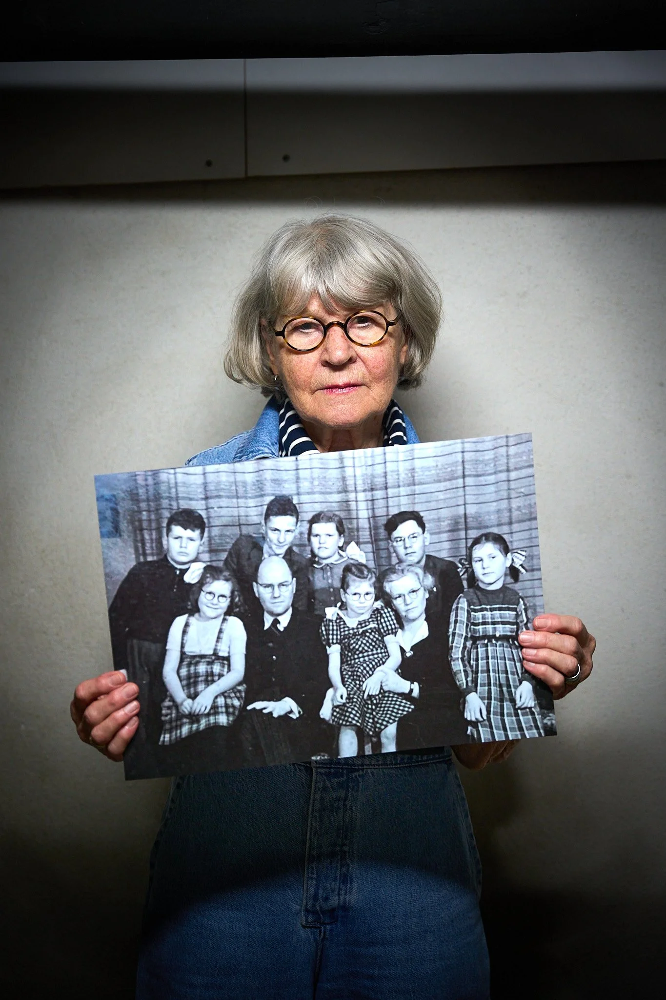An elderly woman with gray hair and glasses holding a vintage black-and-white family photograph with multiple people, some children, seated in front of a curtain.