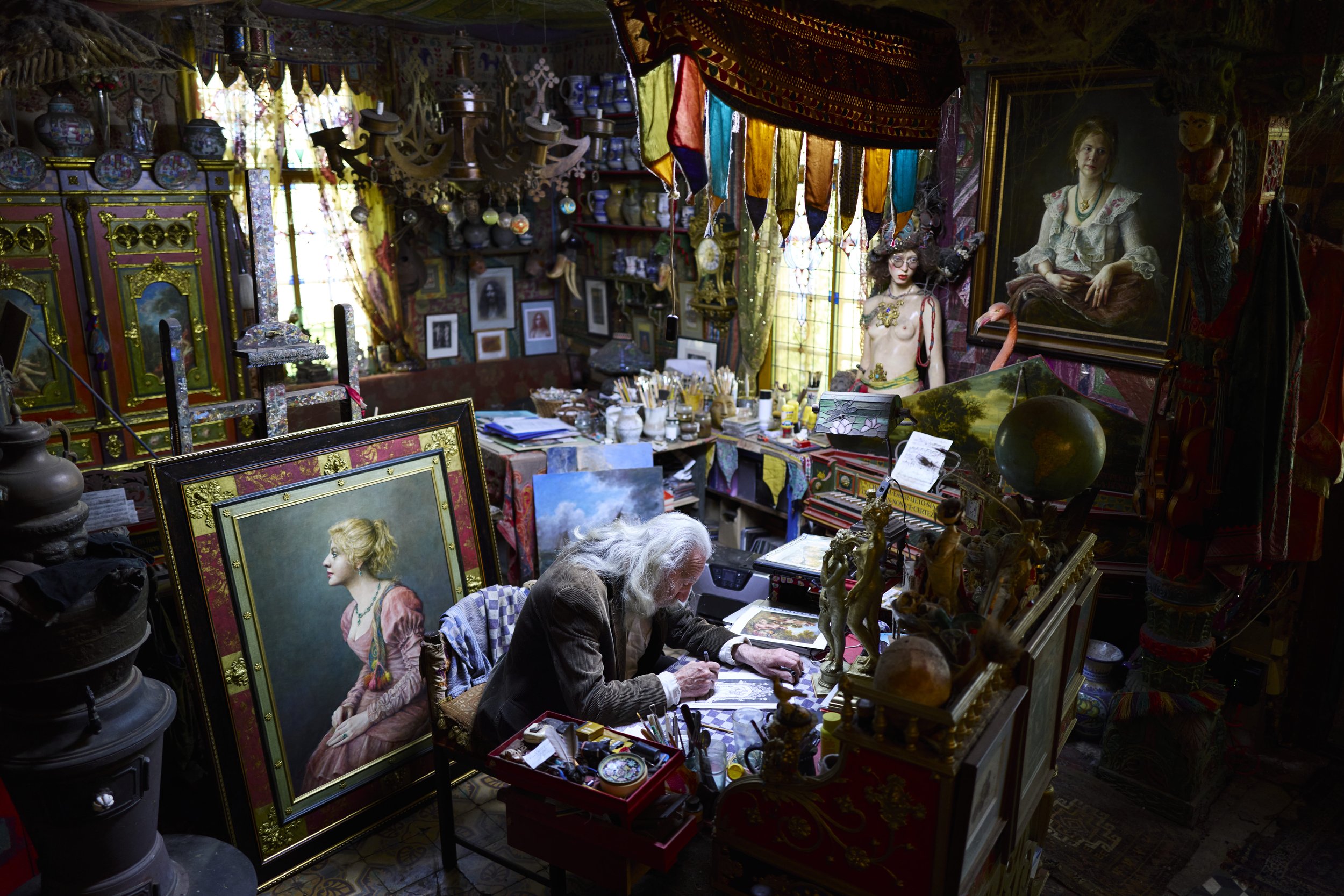 An elderly man with long white hair and a beard, wearing a brown jacket and white shirt, seated at a cluttered desk in an artist's studio filled with paintings, sculptures, and decorative objects.