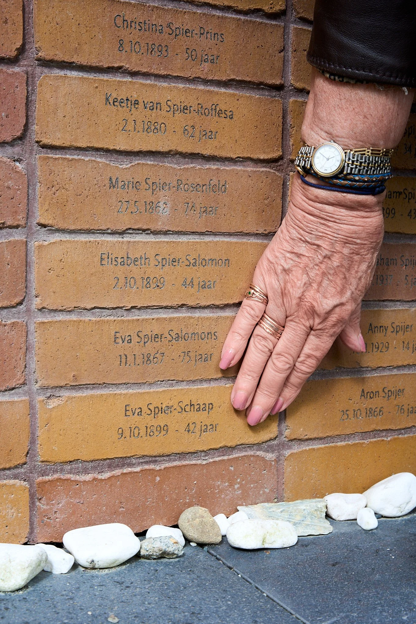 A person's hand resting on a brick memorial wall with engraved names and dates. The wall features names, birth dates, and ages of individuals, with some stones placed at the base of the wall.