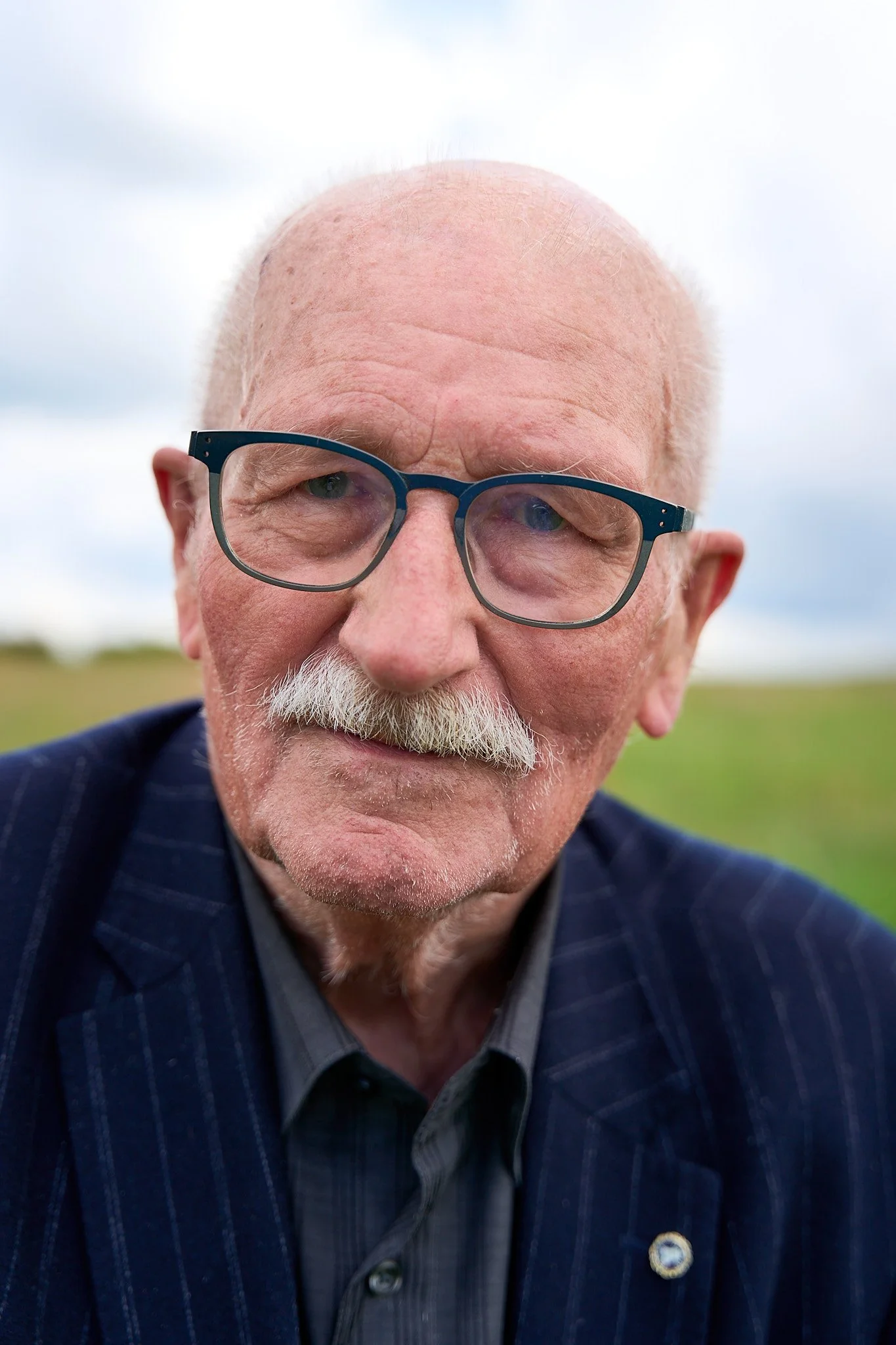 Close-up of an elderly man with glasses, a mustache, and a beard, wearing a dark blazer and a checkered shirt, outdoors with a cloudy sky in the background.