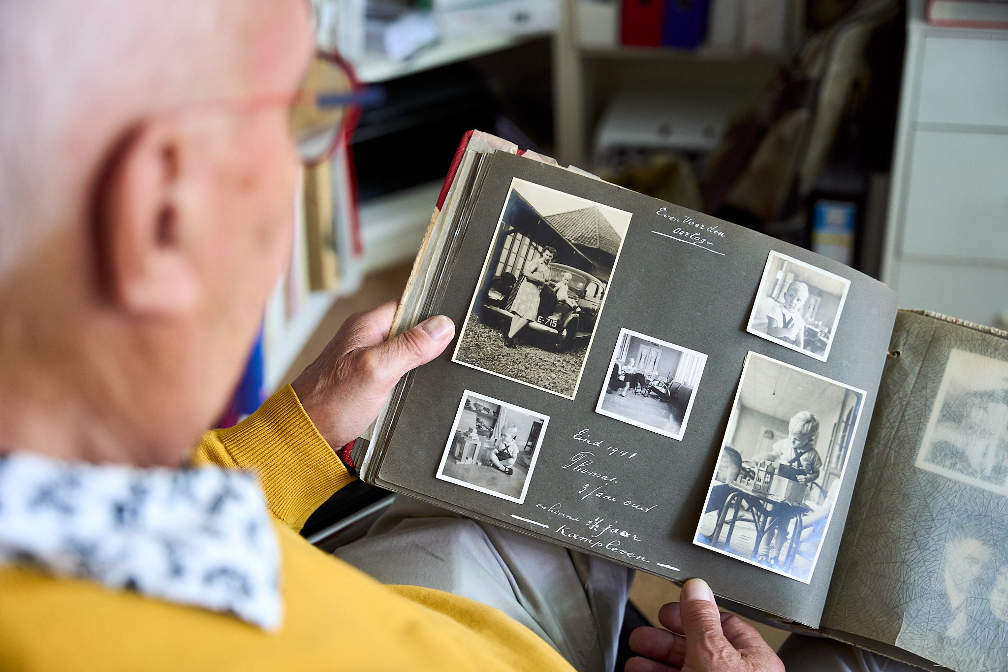An elderly person wearing glasses and a yellow sweater looking at a scrapbook with black-and-white photos of a woman and children, with handwritten notes.