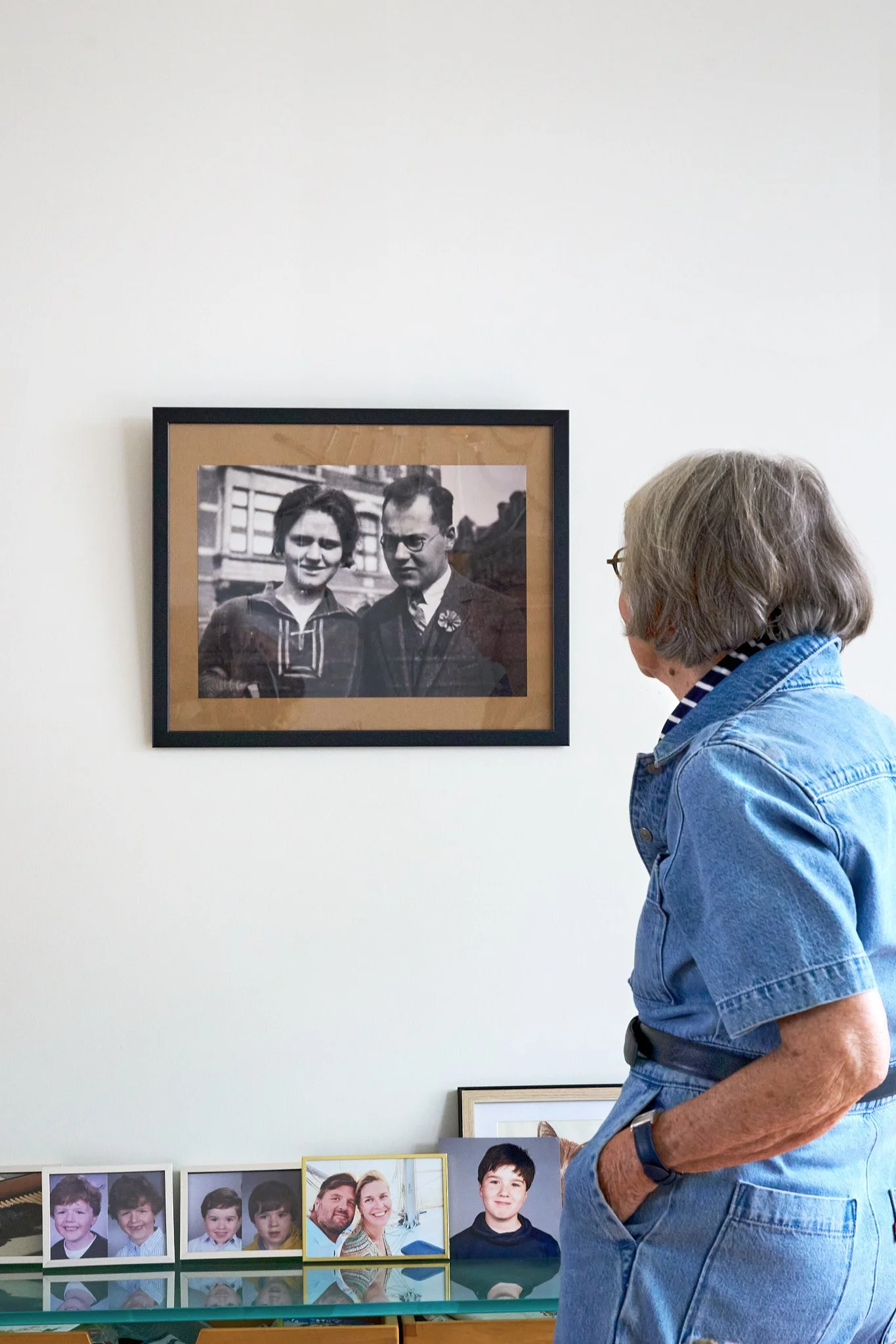 An elderly woman looking at a framed black-and-white family photograph on a wall. The woman is wearing a denim dress and glasses. Below on a table, there are several smaller, colorful family photographs.