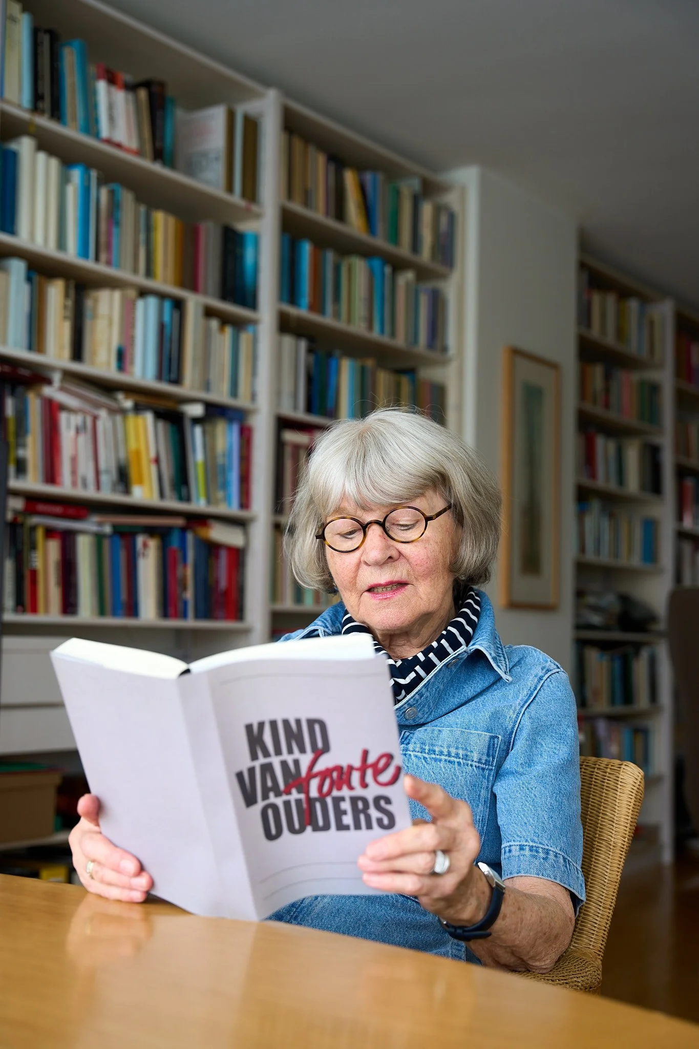 An elderly woman with gray hair and glasses, wearing a denim shirt and striped scarf, reading a book titled 'KIND VAND OUERS' at a wooden table in a room filled with bookshelves.