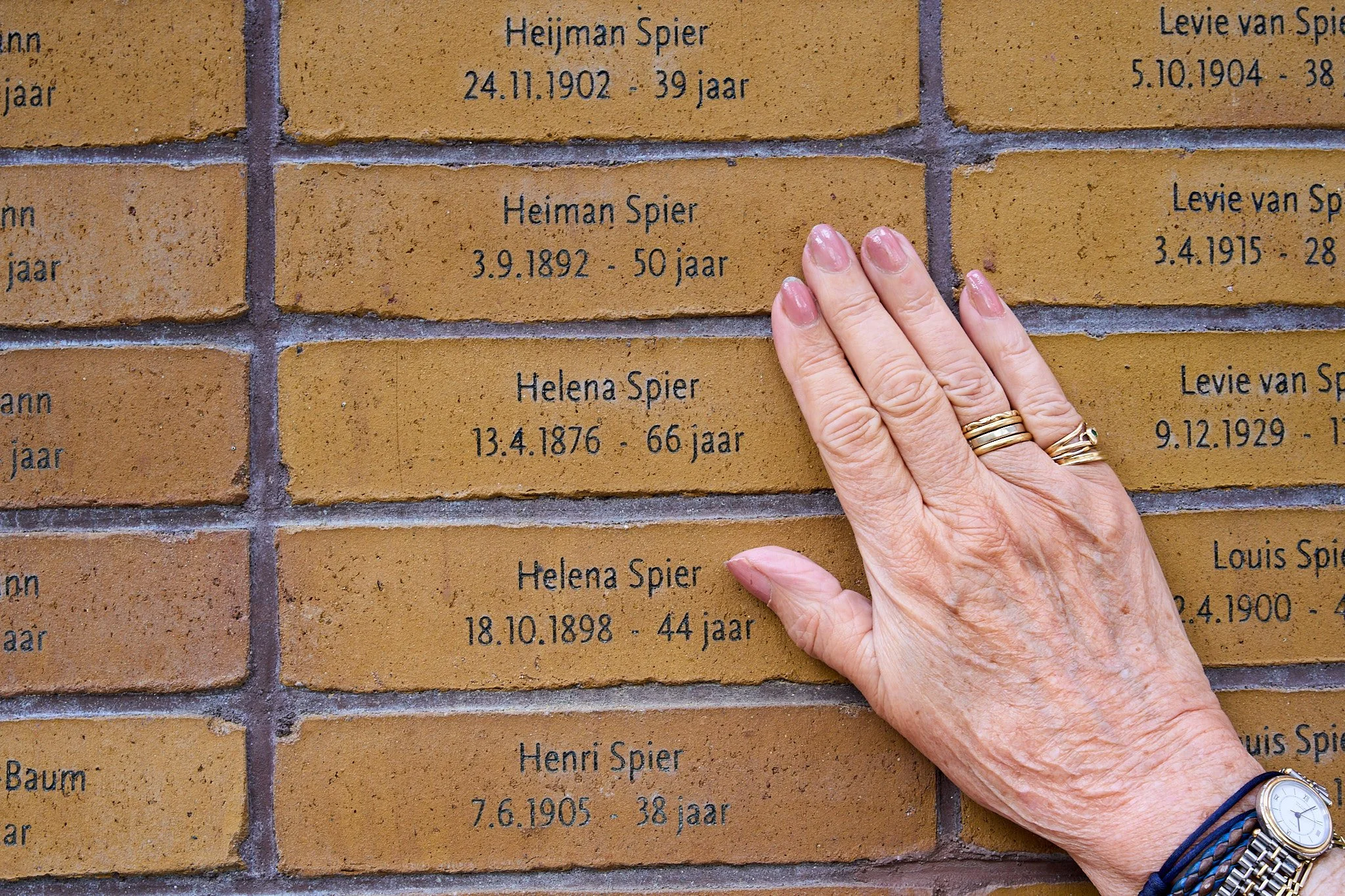 Close-up of a hand with rings and a watch touching a brick wall with engraved names and birth and death dates.