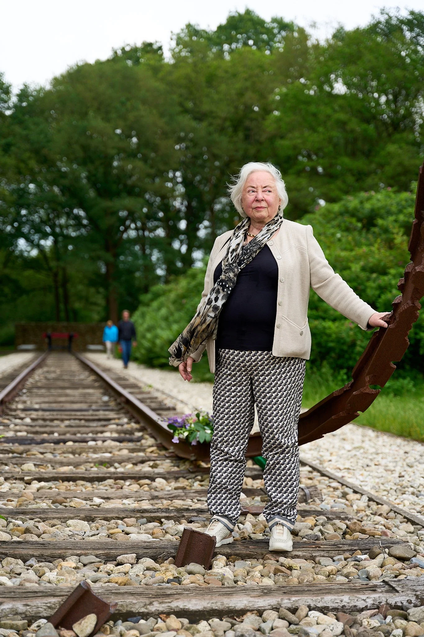 An elderly woman with white hair standing on an abandoned railroad track, holding a large rusted gear, with two people walking in the background.