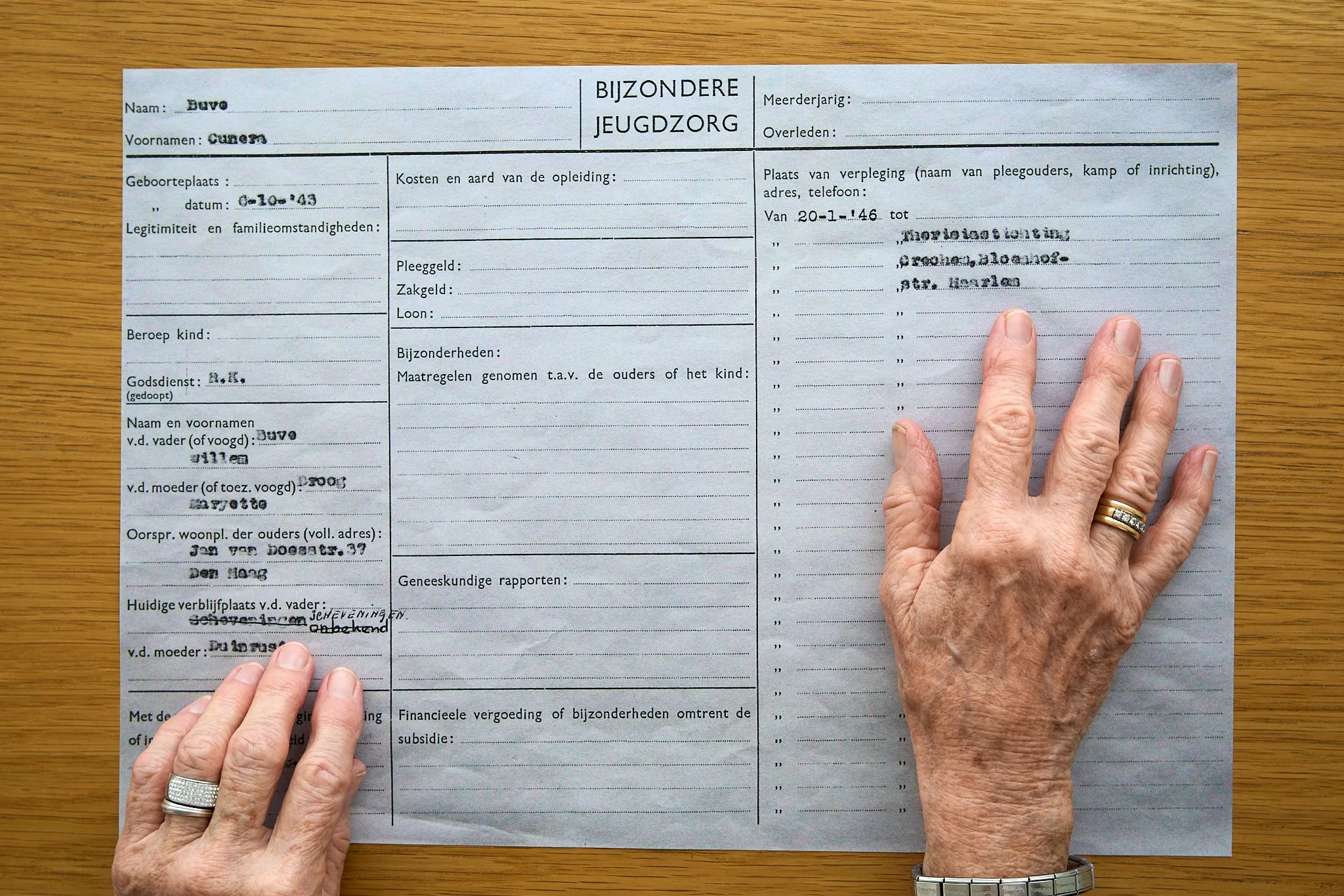 A person with aged hands, placed on a document, pointing at a section. The document is a form with handwritten and typed text, including various fields filled in, laid on a wooden surface.