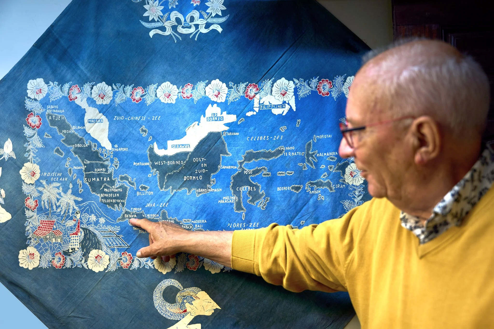 A man wearing glasses and a yellow sweater points to a map of Southeast Asia, which features islands and countries like Singapore, Borneo, and the Philippines, decorated with floral patterns and figures.