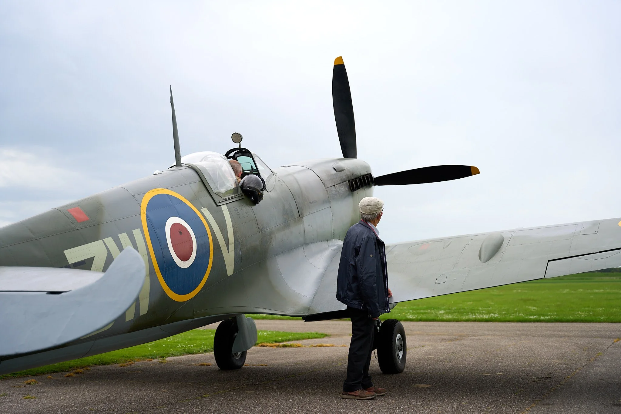A vintage fighter aircraft with the marking of a Royal Air Force roundel is parked on the tarmac with an older man standing nearby, both on an overcast day.