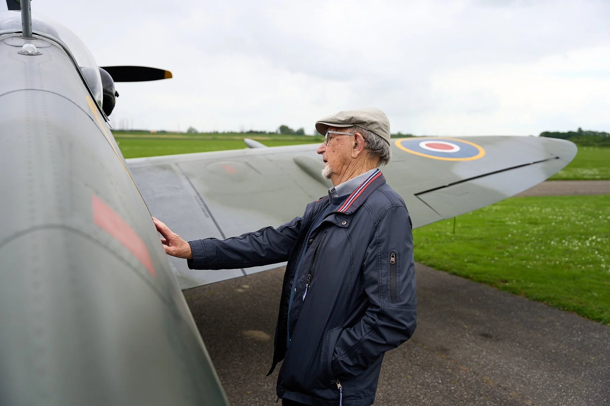 An elderly man in a navy jacket and beige cap standing beside a vintage fighter airplane on a grassy airfield.