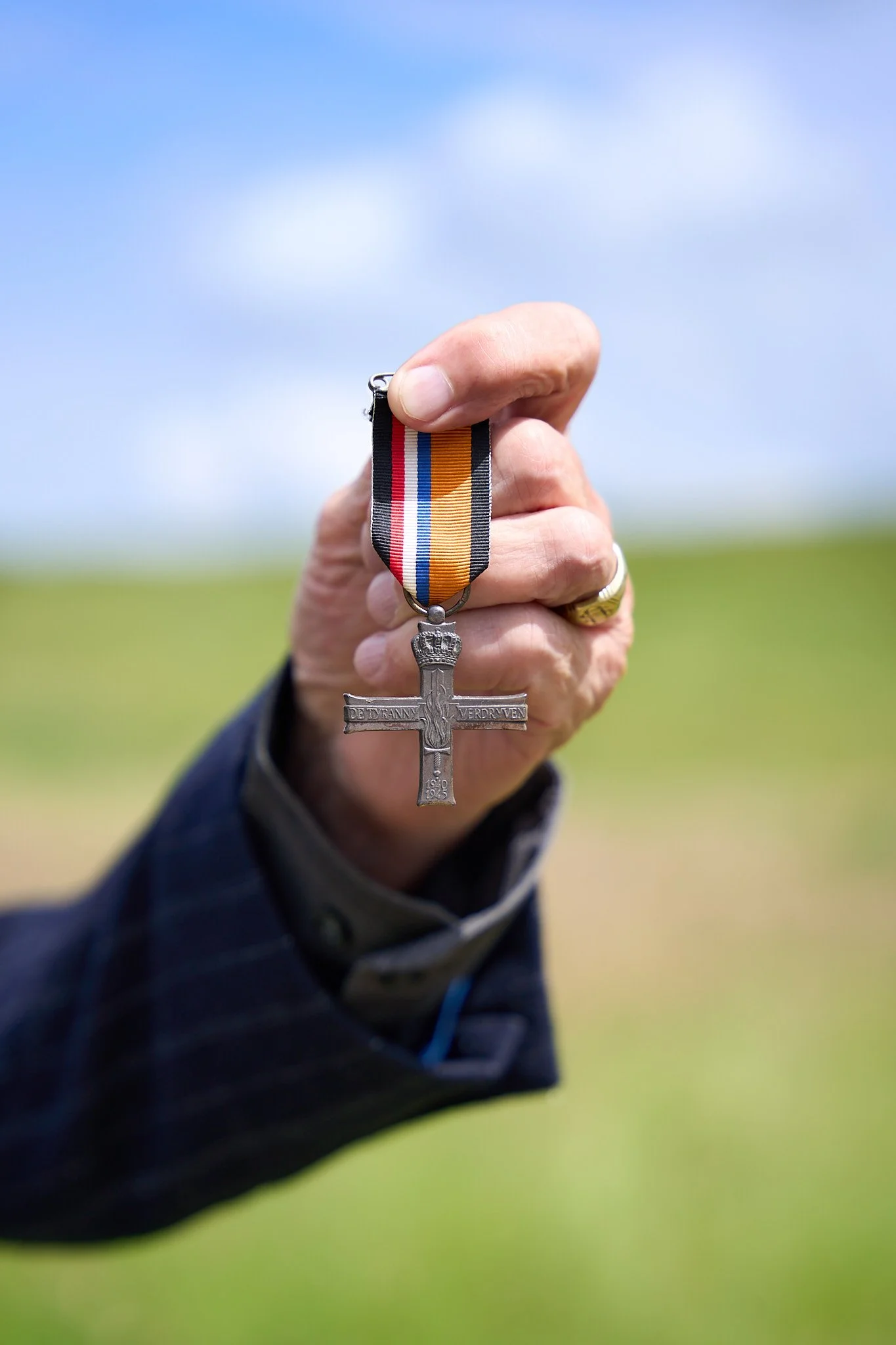 Close-up of a hand holding a medal with a crucifix pendant against a blue sky and green field background