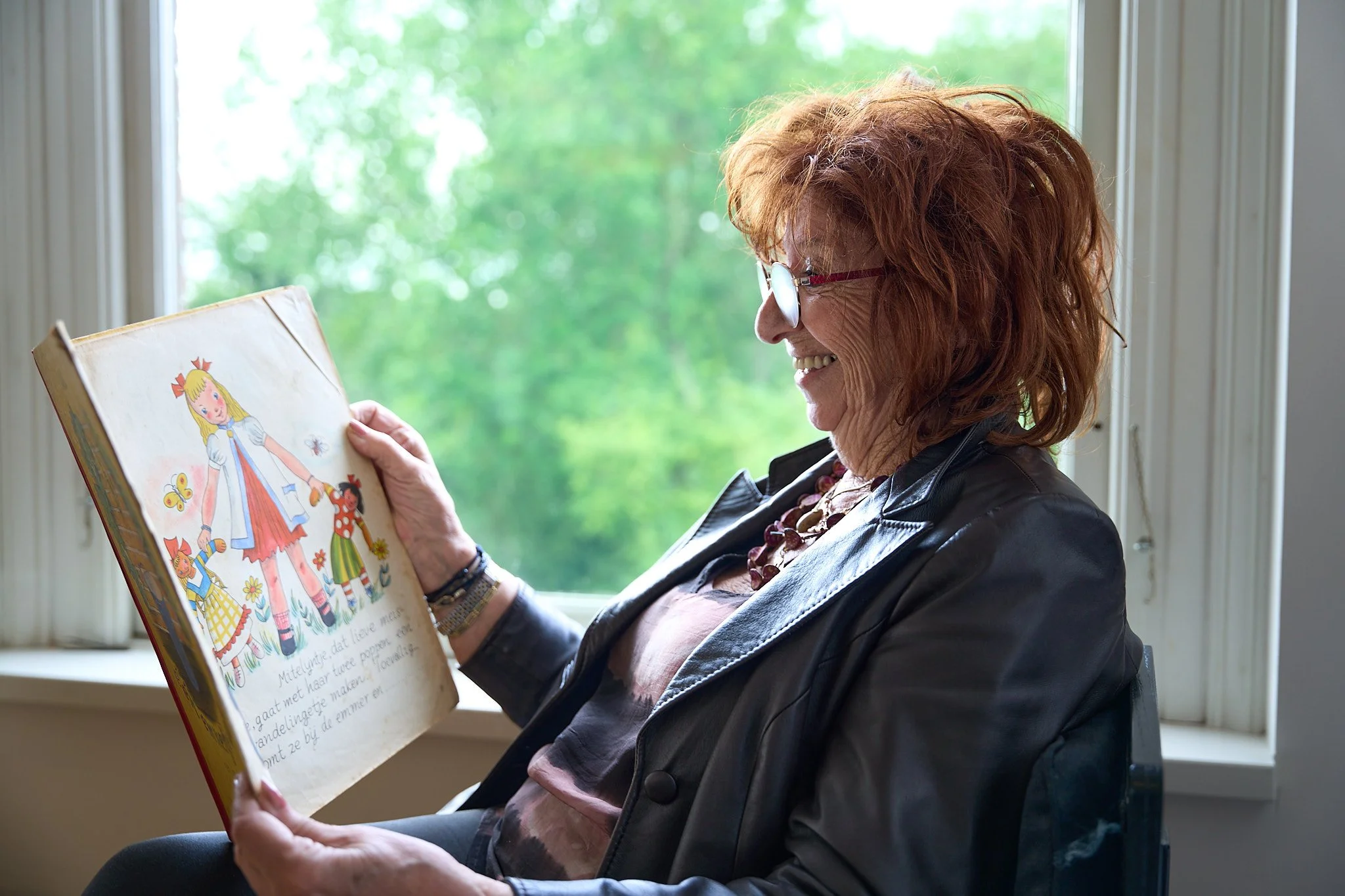 A woman with red hair, glasses, and a leather jacket reading a children's picture book with an illustration of a princess at a window.