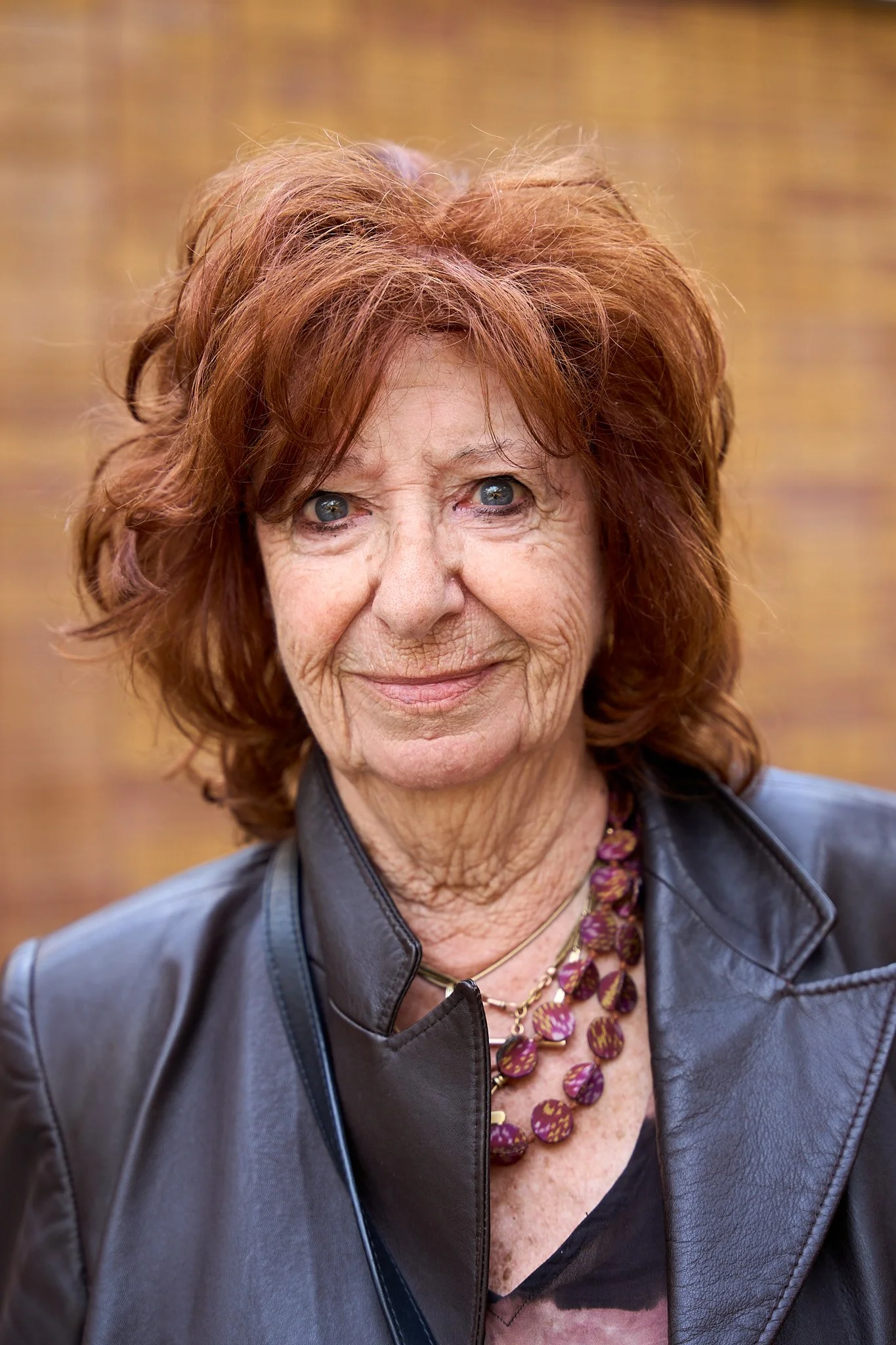 A portrait of an elderly woman with curly red hair, wearing a leather jacket and a purple beaded necklace, smiling against a blurred brick wall background.