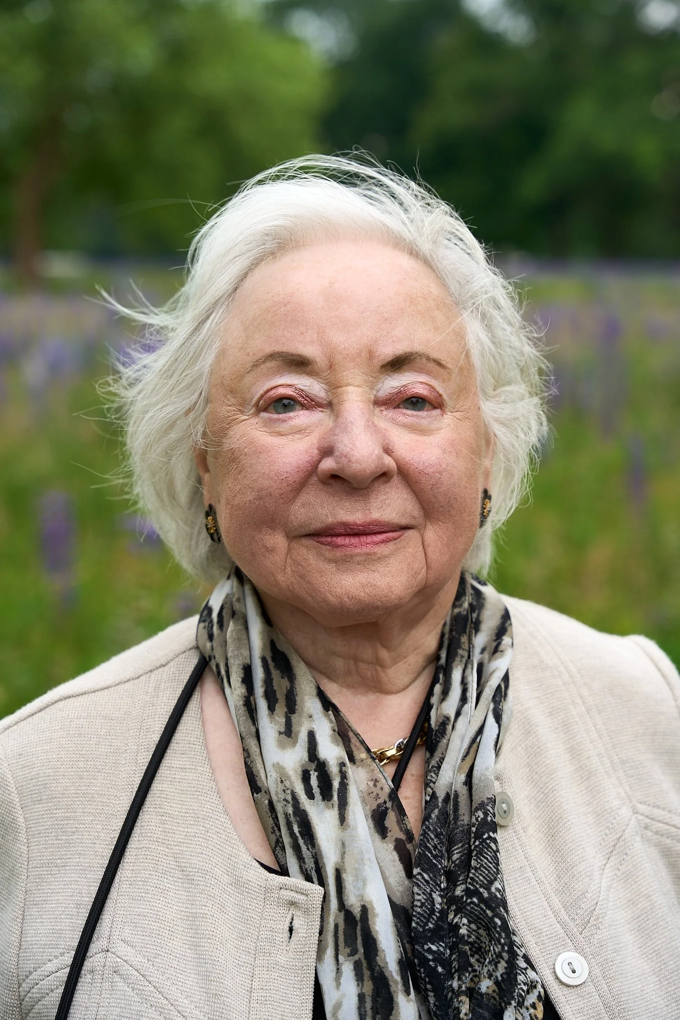 An elderly woman with white hair standing outdoors in a field of flowers, wearing a beige jacket, a patterned scarf, and earrings.