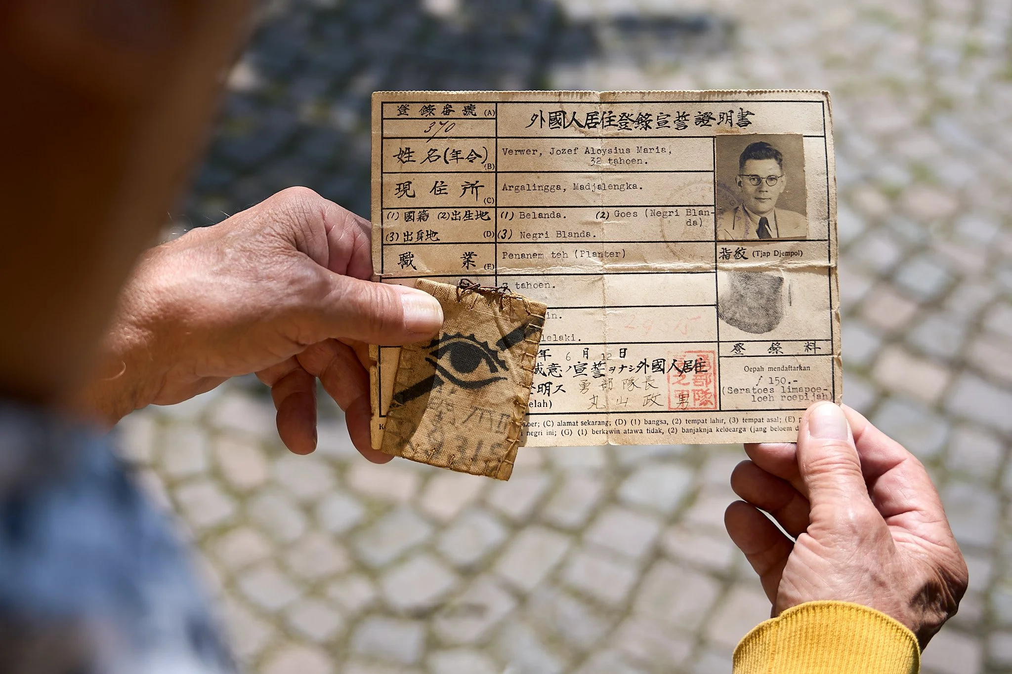 Person holding old Indonesian identification card with a photo, handwritten details, and red stamps, outdoors on cobblestone ground.