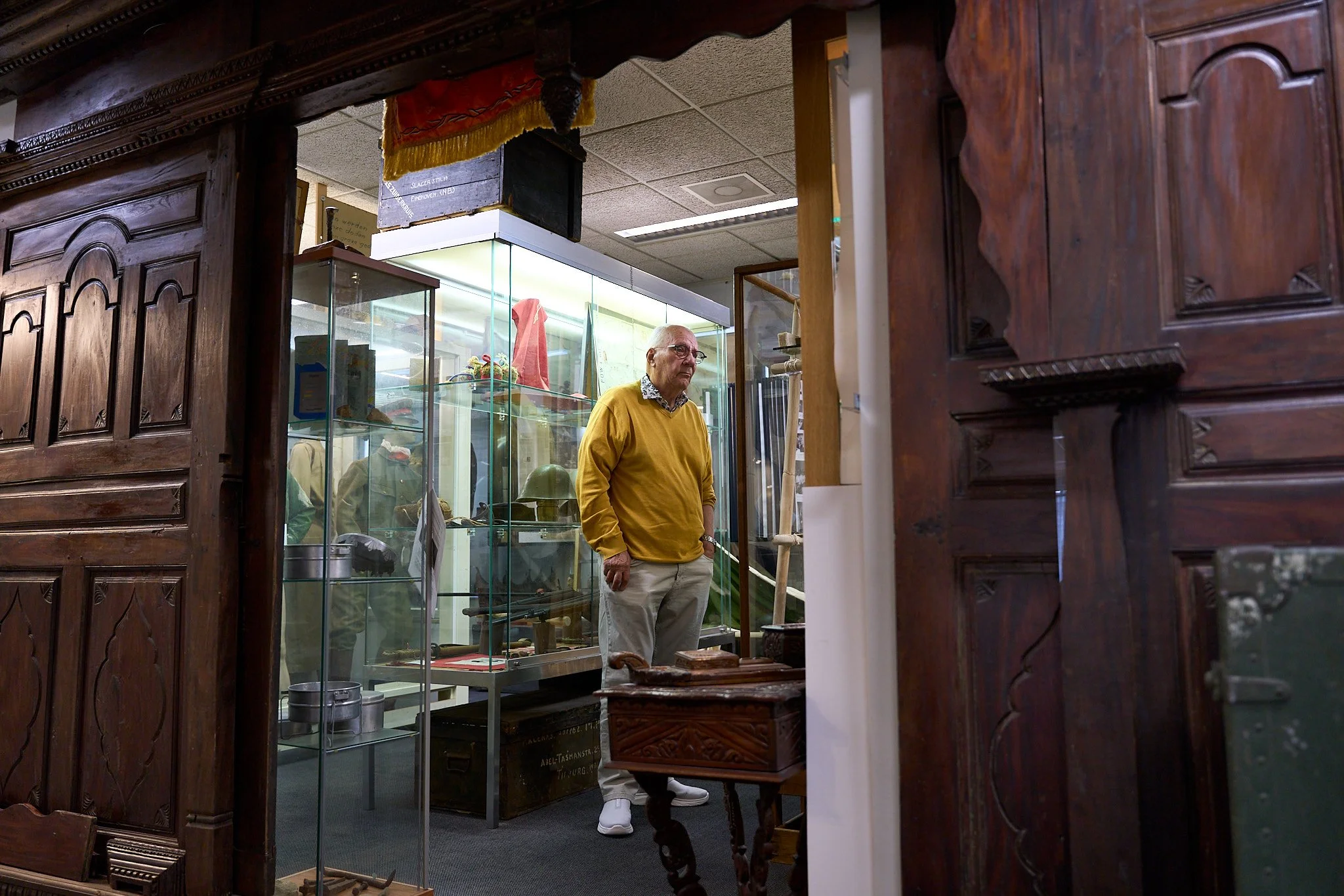 An elderly man in a yellow sweater and white pants standing inside a museum exhibit, viewed through an open wooden doorway with glass display cases and mannequins dressed in military uniforms in the background.