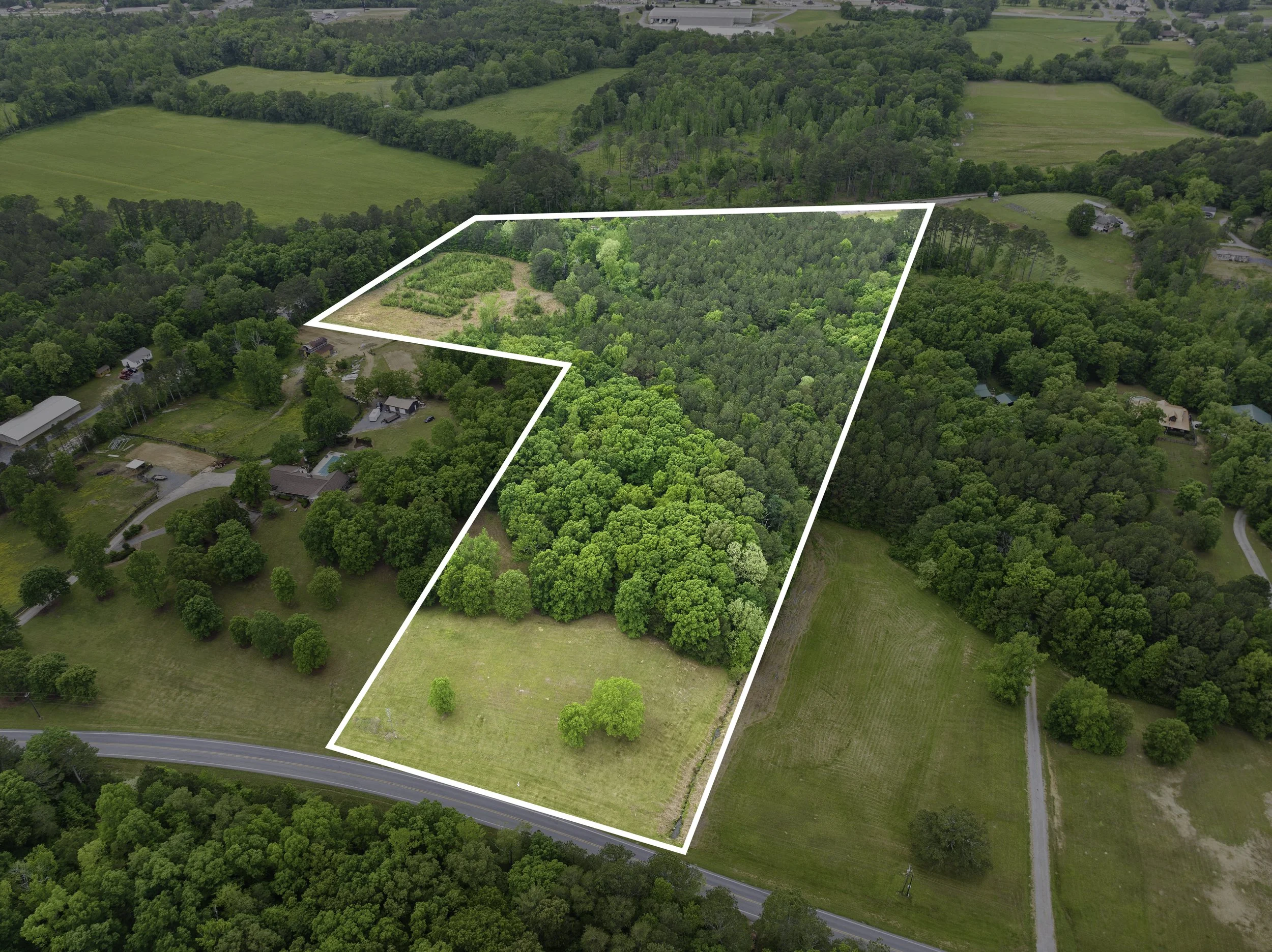 Aerial view of a large green forested property with a white boundary outline, adjacent to residential homes and rural farmland, with a paved road at the bottom edge.