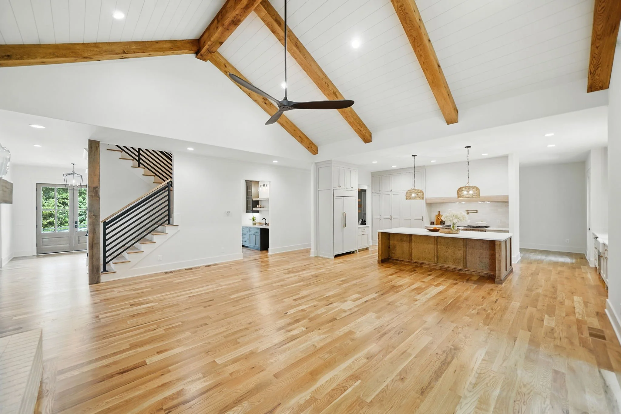 Spacious open-plan living area with wooden floors, white walls, a vaulted ceiling with exposed wooden beams, a black ceiling fan, and a modern kitchen with a large island, white cabinets, and pendant lighting.