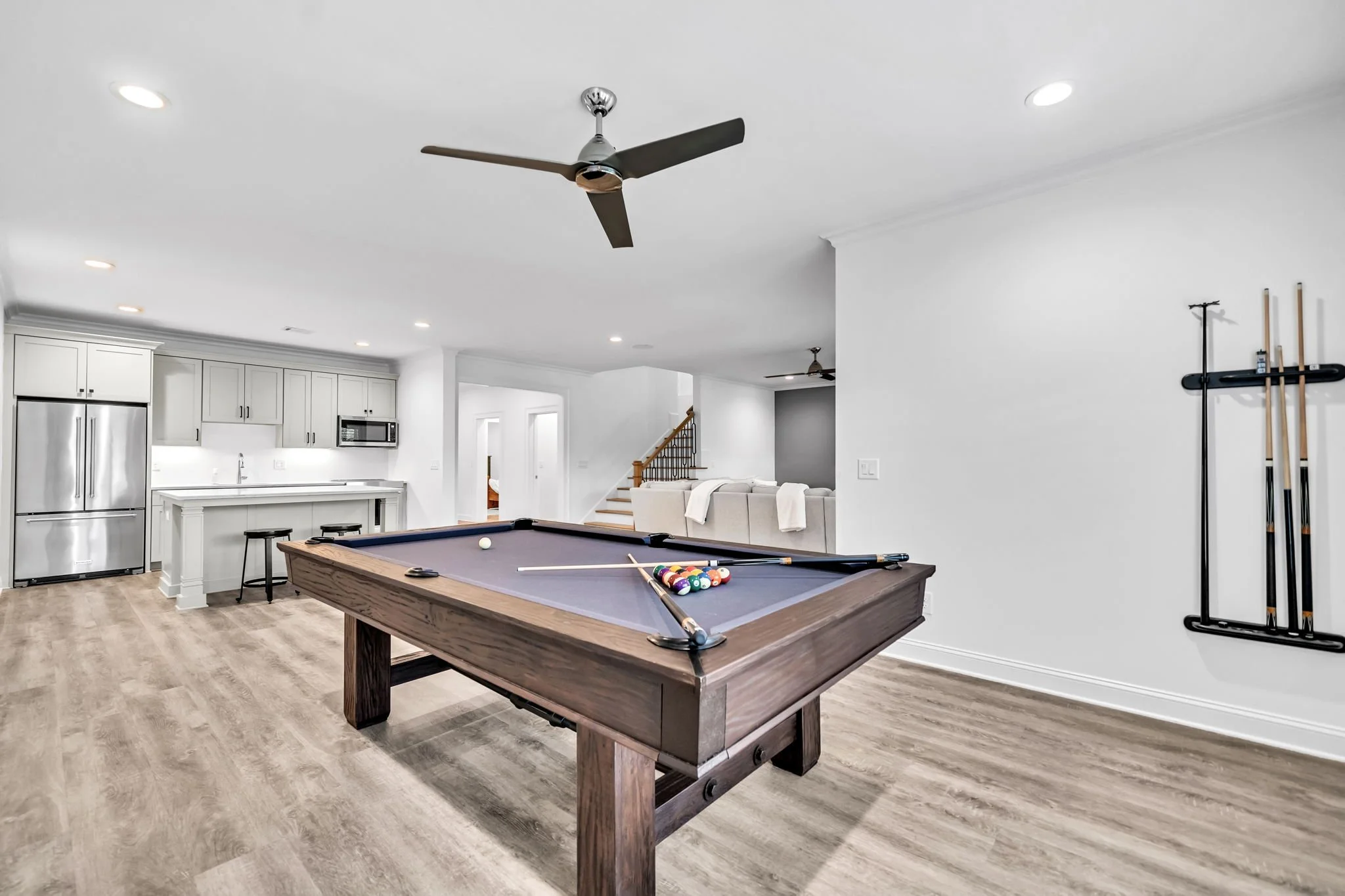 Living room with a pool table, kitchen area with white cabinets and a refrigerator, wall-mounted pool cue rack, ceiling fans, and a staircase.