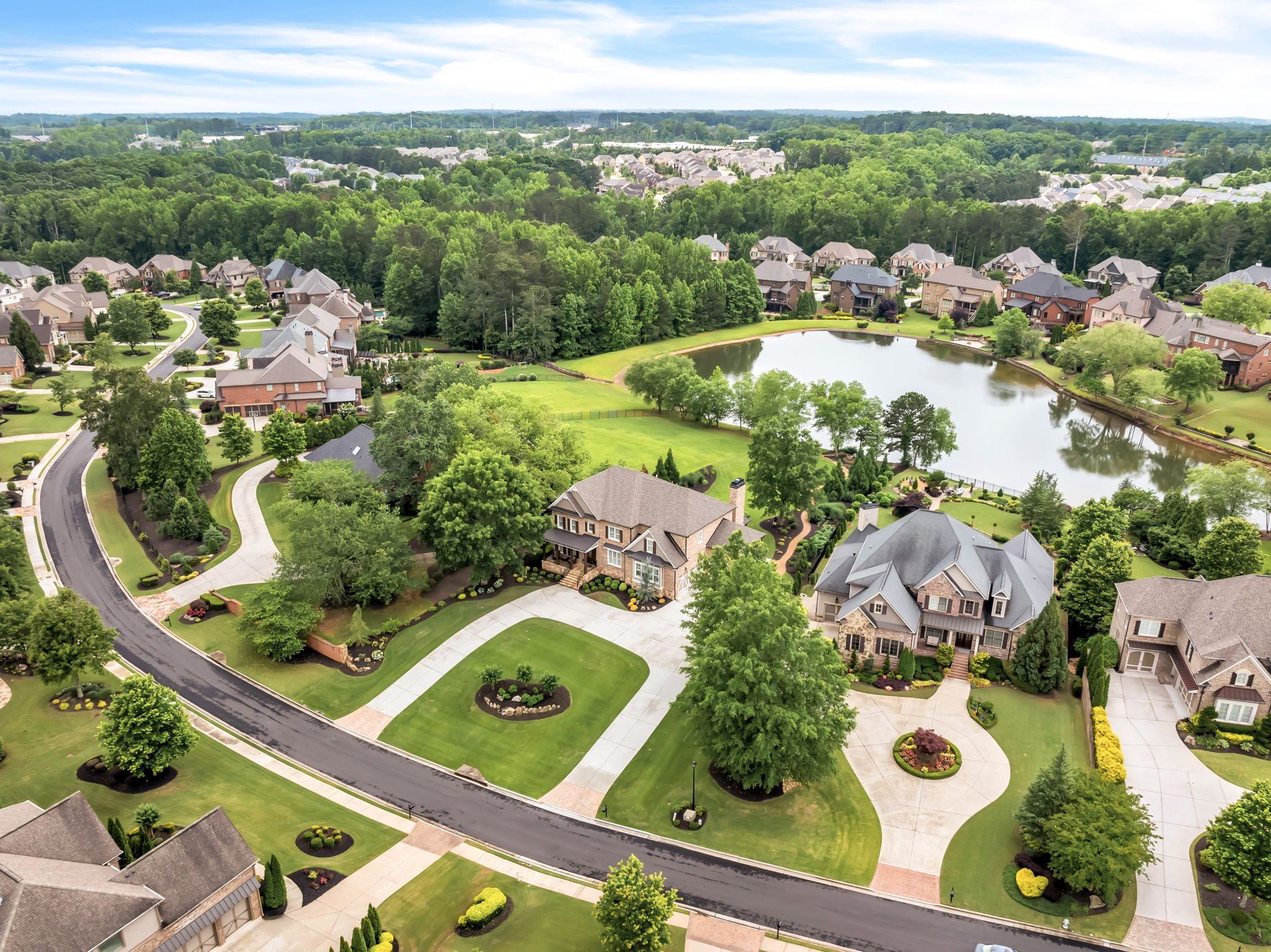 Aerial view of a suburban neighborhood with large houses, green lawns, trees, a lake, and winding roads.