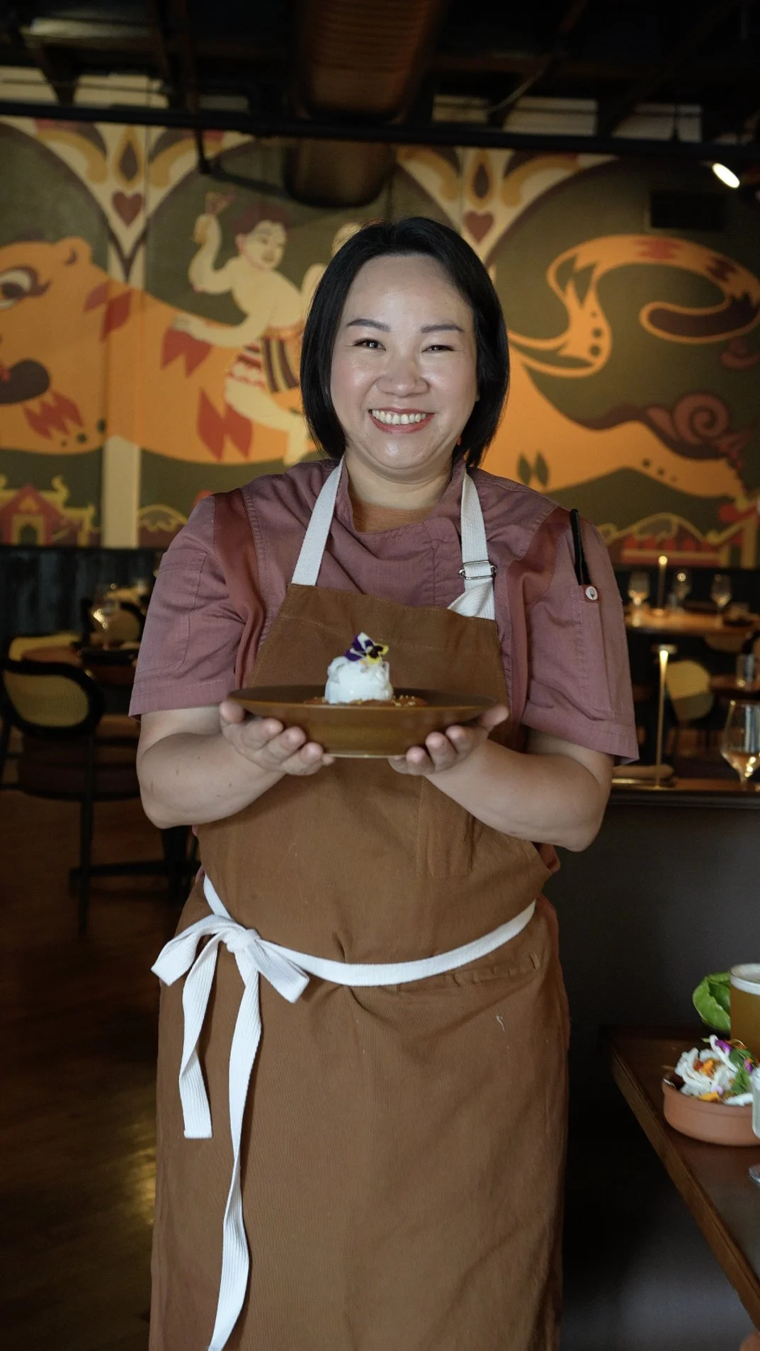 A smiling woman in a pink shirt and brown apron holding a plate with a dessert in a restaurant with a colorful mural in the background.