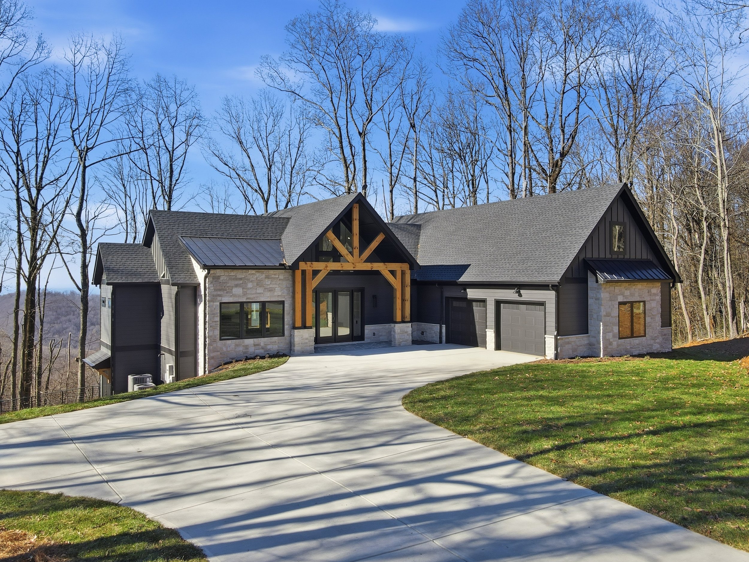 Modern house with stone and dark siding exterior, pitched roof, black garage doors, wooden accents, large windows, and a curved concrete driveway surrounded by a grassy lawn and leafless trees under a blue sky.