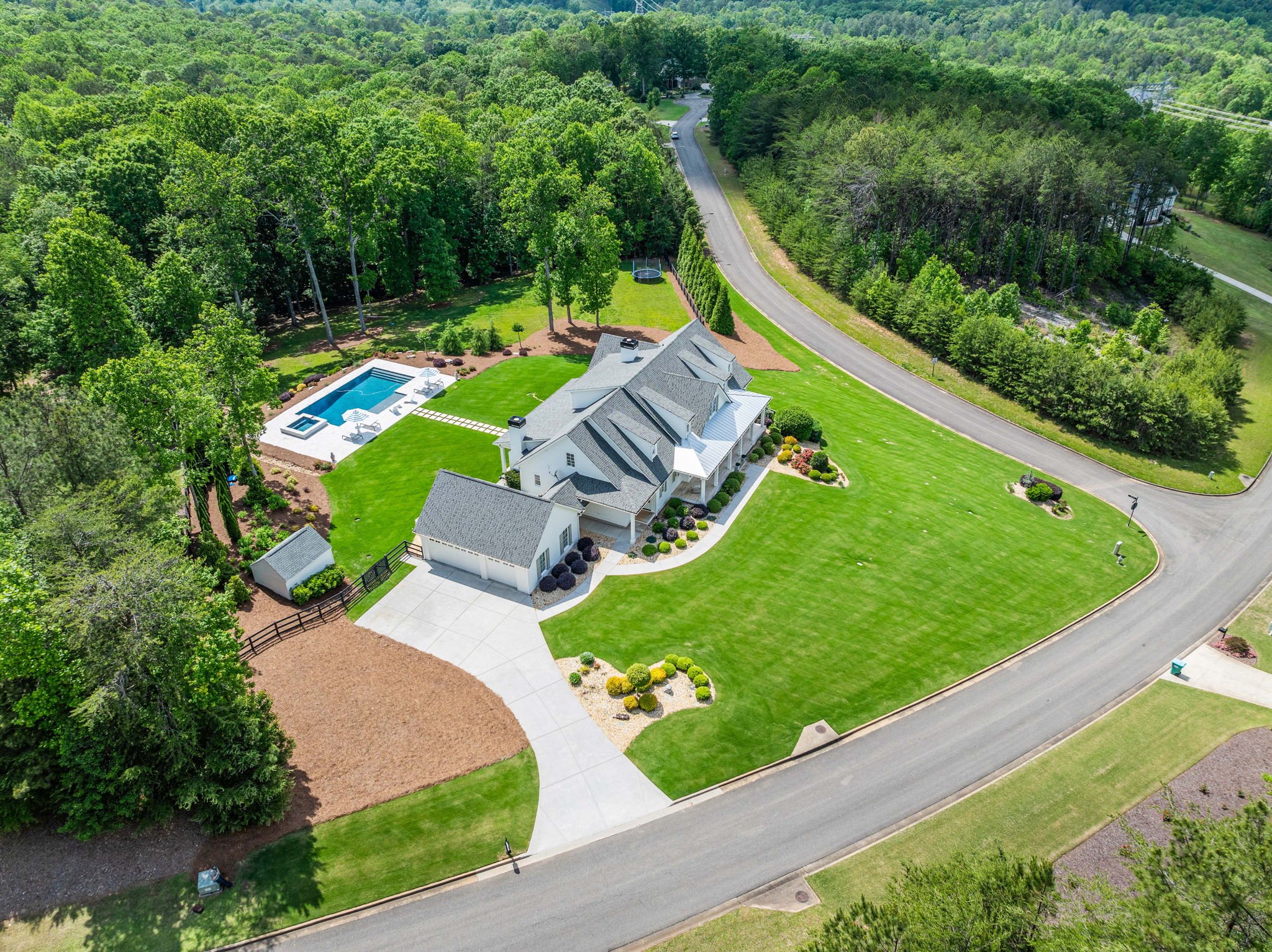 A large house with a gray roof and white exterior walls, surrounded by a well-manicured lawn, with a paved driveway leading to a garage, a backyard with a swimming pool, surrounded by trees, and a winding road nearby.