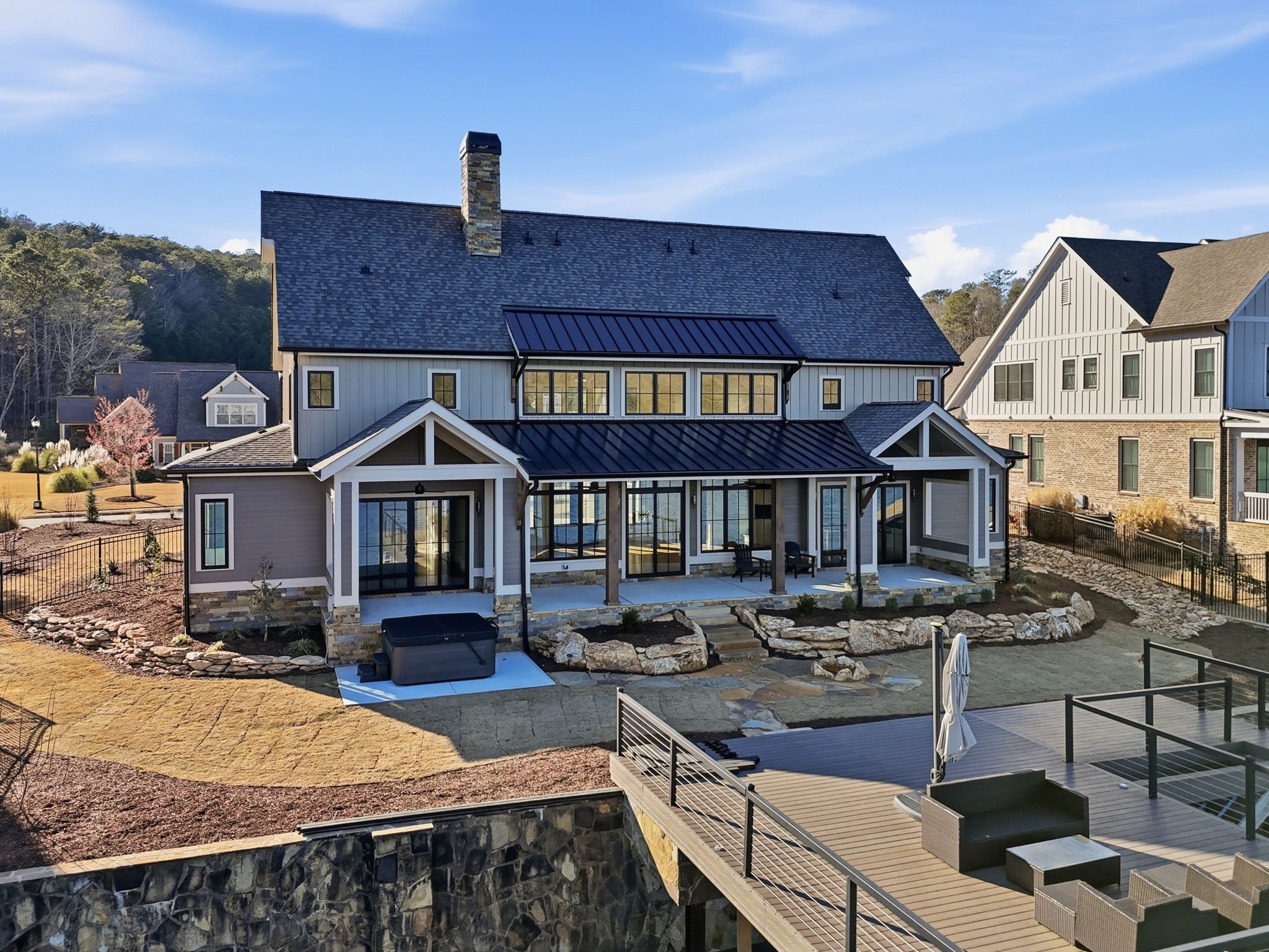 A modern two-story house with a stone and siding exterior, large windows, a metal roof, and a covered porch with seating. The backyard features a deck, a hot tub, landscaping with rocks and grass, and surrounding neighboring homes.