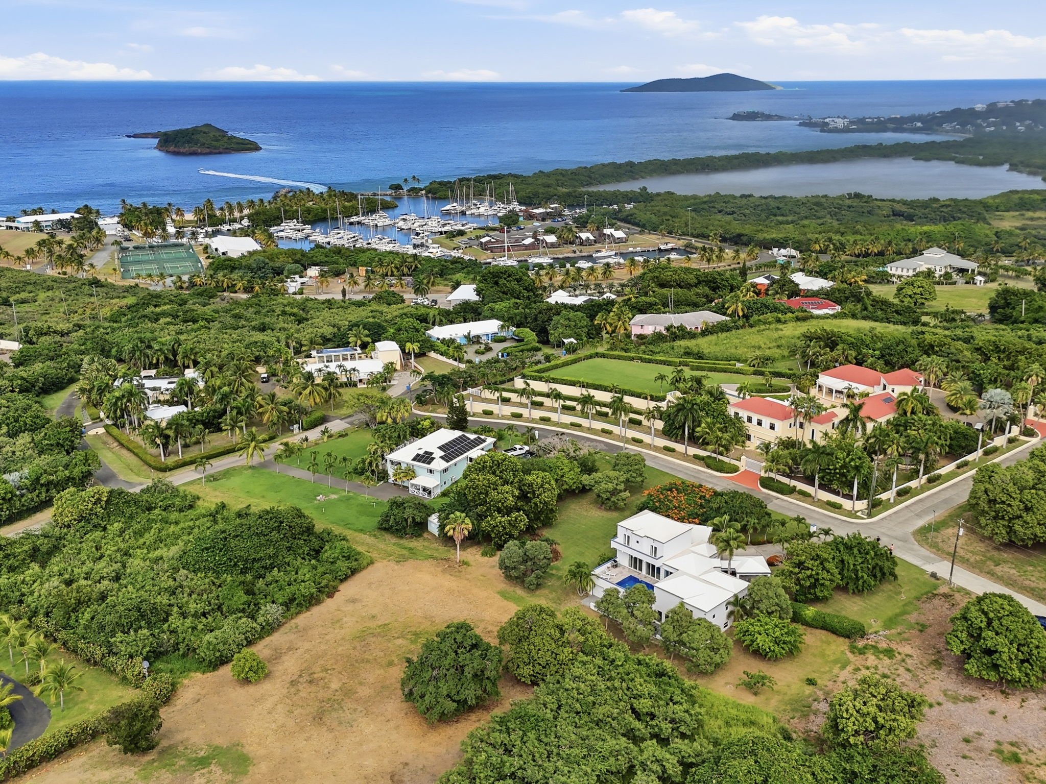 Aerial view of a coastal residential area with green lawns, trees, houses with red and white roofs, surrounded by lush vegetation, near a marina with boats, and the ocean in the background with islands and a cloudy sky.