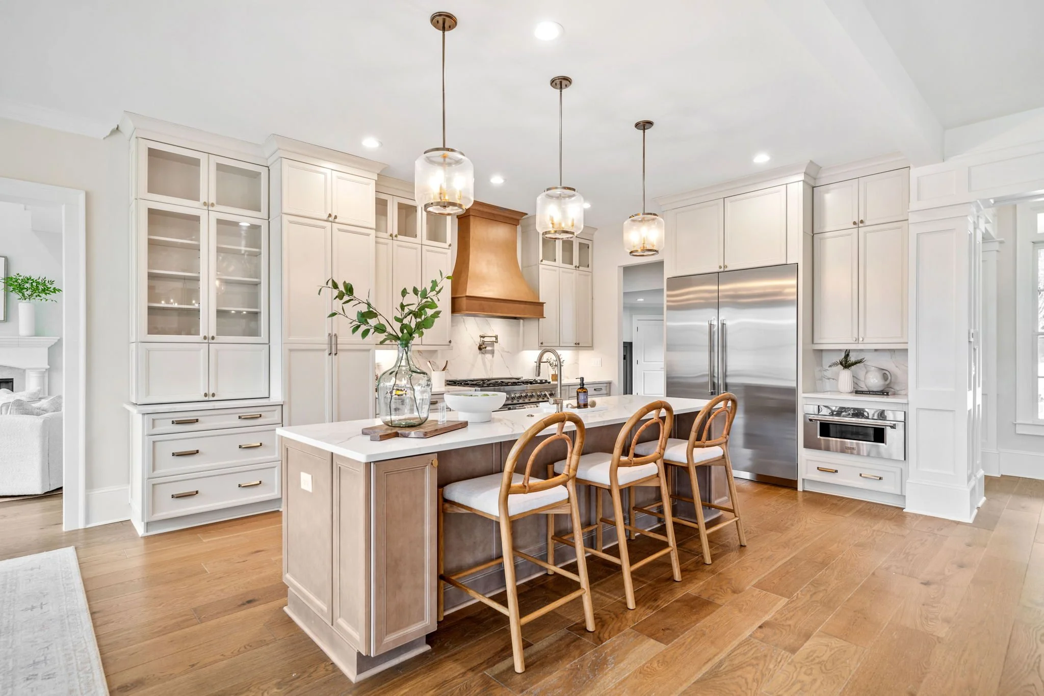 Modern kitchen with white cabinets, stainless steel refrigerator, wooden floor, island with seating, pendant lights, and a cooktop with copper range hood.