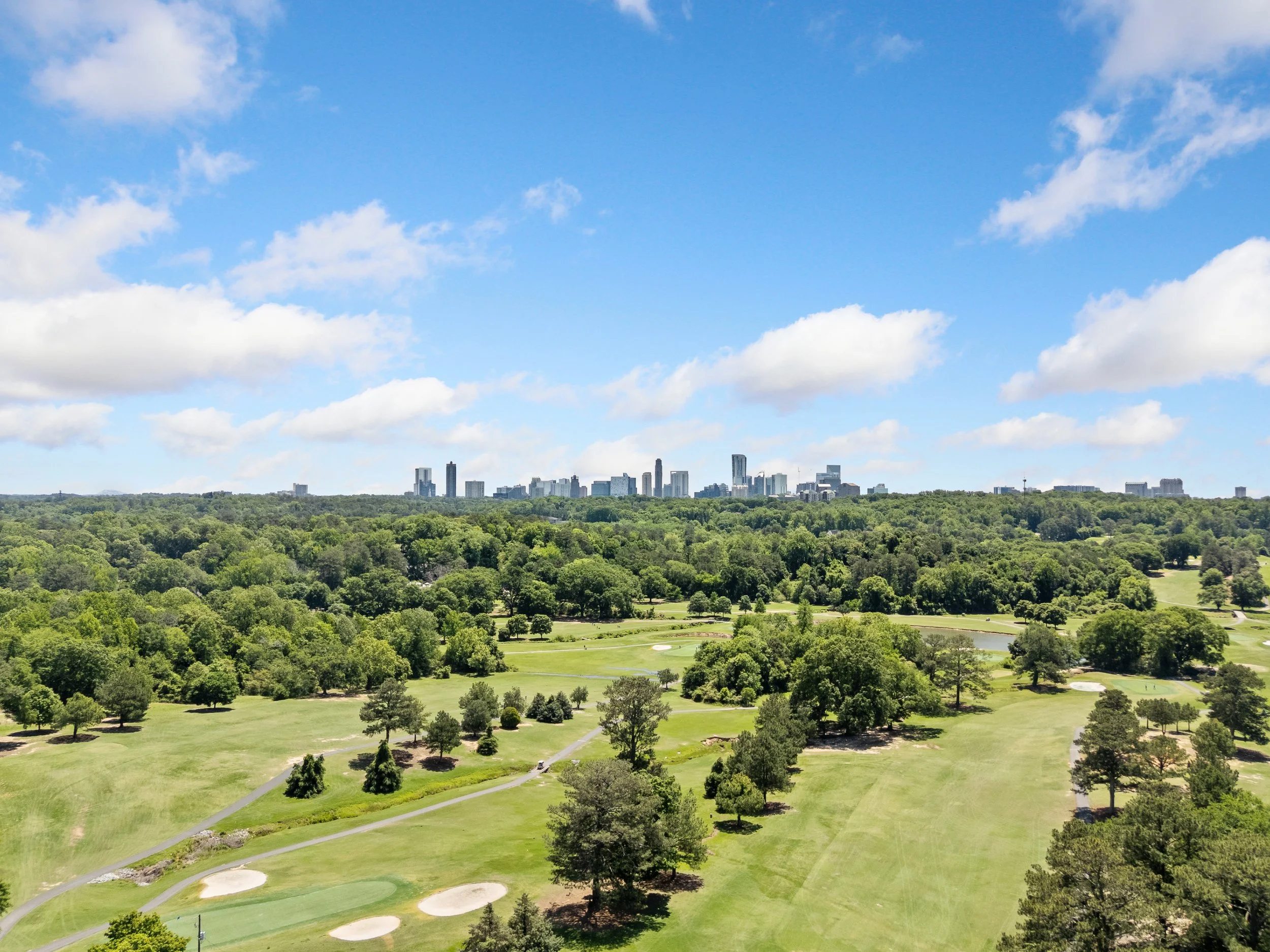 A panoramic view of a golf course with green fairways and sand traps, surrounded by trees, with a city skyline with tall buildings in the background under a partly cloudy sky.