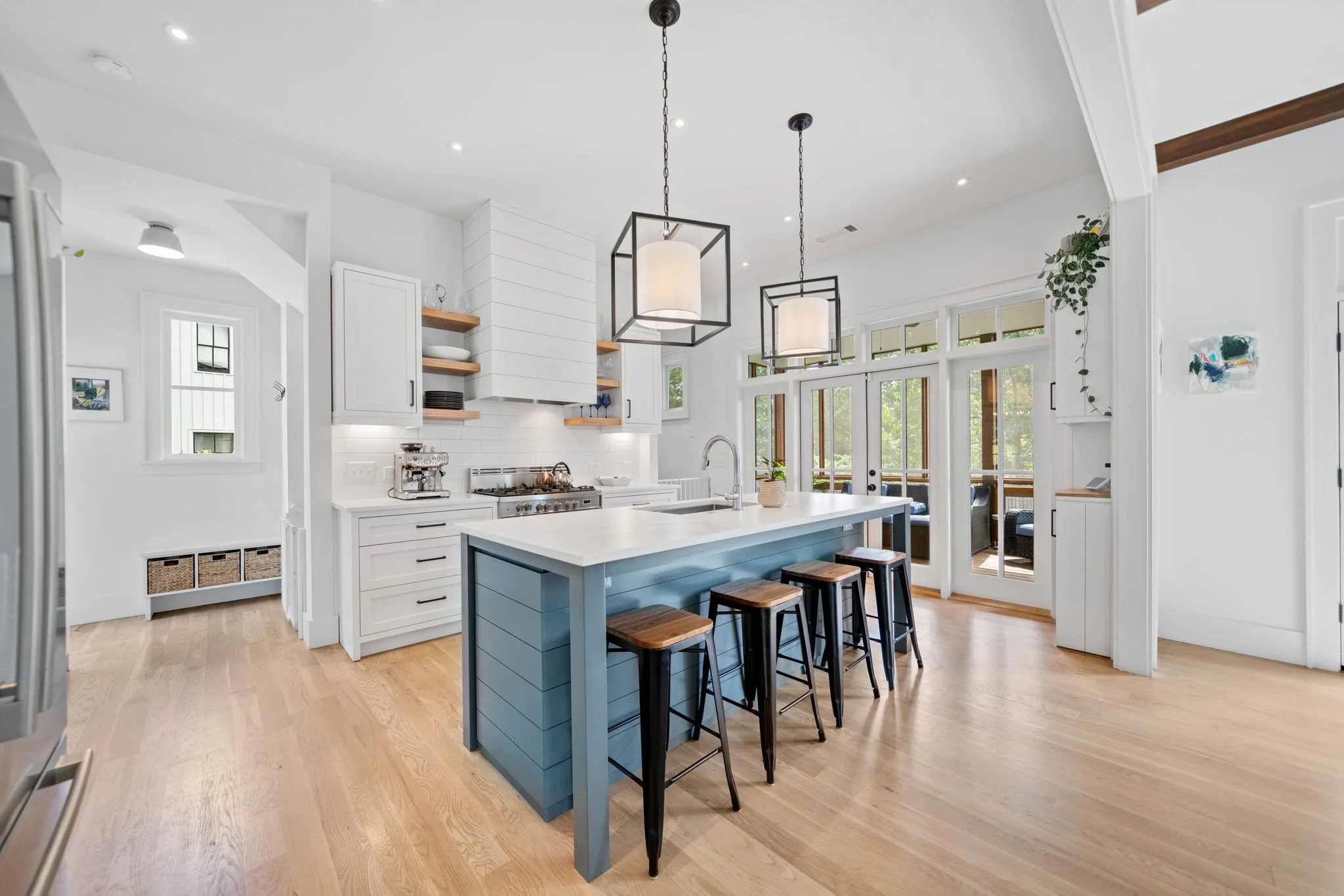Modern kitchen with white cabinets, a large island with a blue base, black stools, open wooden shelves, and pendant lights. French doors lead to a balcony with outdoor seating.