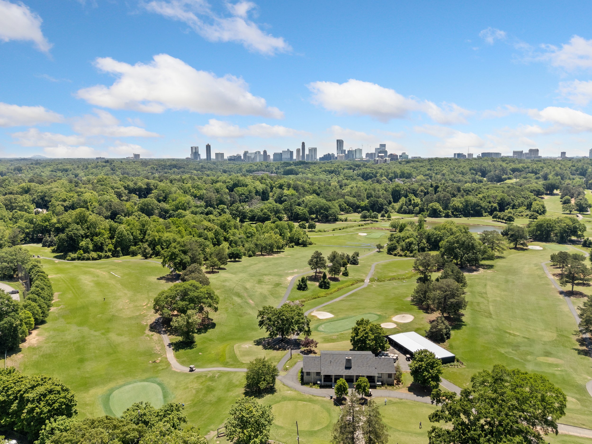 Aerial view of a golf course with green fairways, sand traps, trees, a clubhouse, and a city skyline in the distance under a partly cloudy sky.