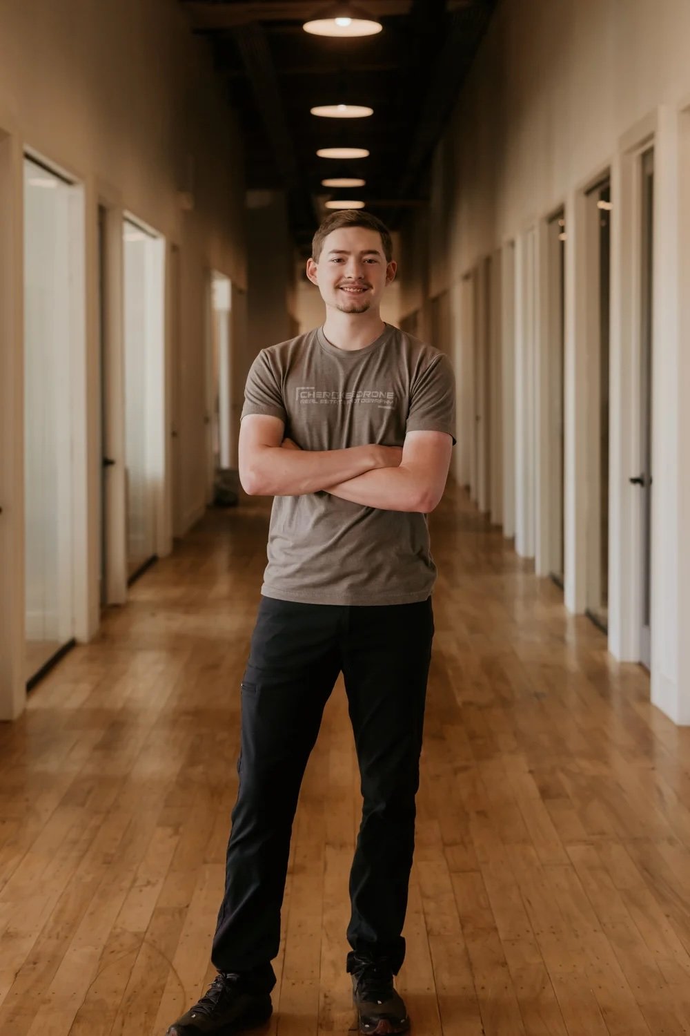 A young man standing with arms crossed, smiling, in a corridor with wooden flooring and multiple doorways.
