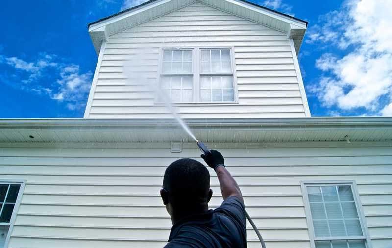 Person pressure washing the exterior of a two-story house with white siding.