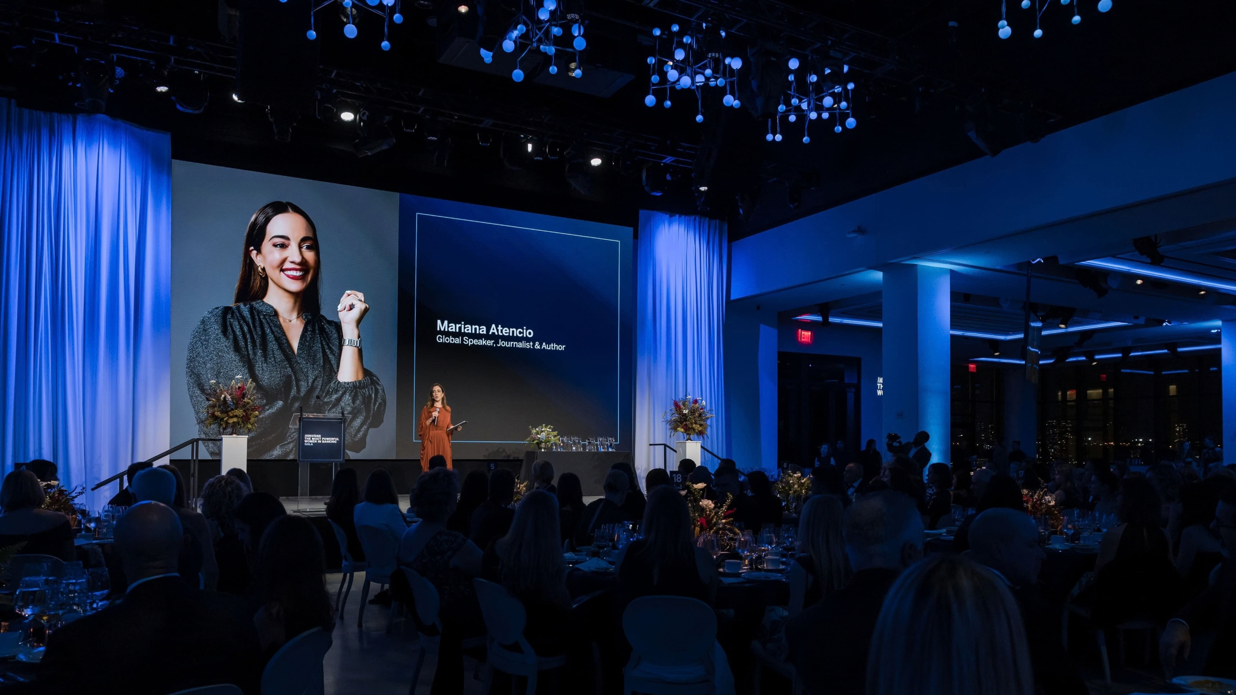 A woman standing on stage at a conference, delivering a speech. The large screen behind her displays her name, Mariana Atencio, and her titles as a global speaker, journalist, and author. The audience is seated in front, with flowers on tables, and the venue features blue curtains and modern lighting fixtures.