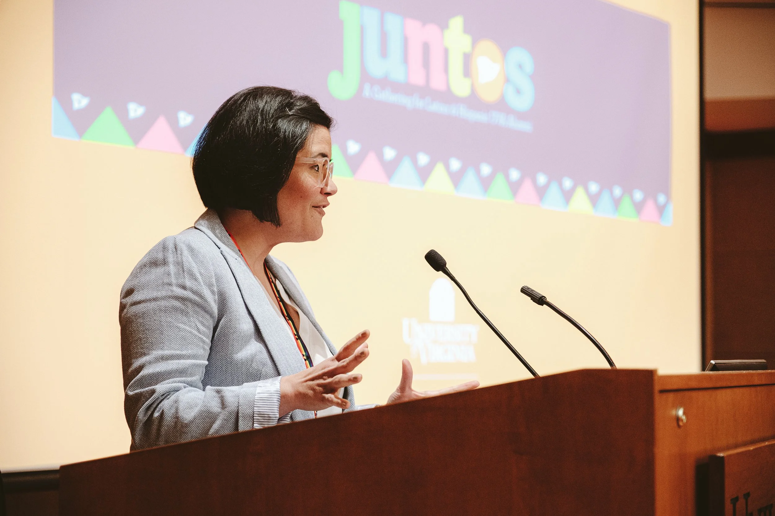 A femme with short layered dark brown hair stands gesticulating at a podium. She is wearing a gray suit jacket and has a rainbow lanyard on her neck.