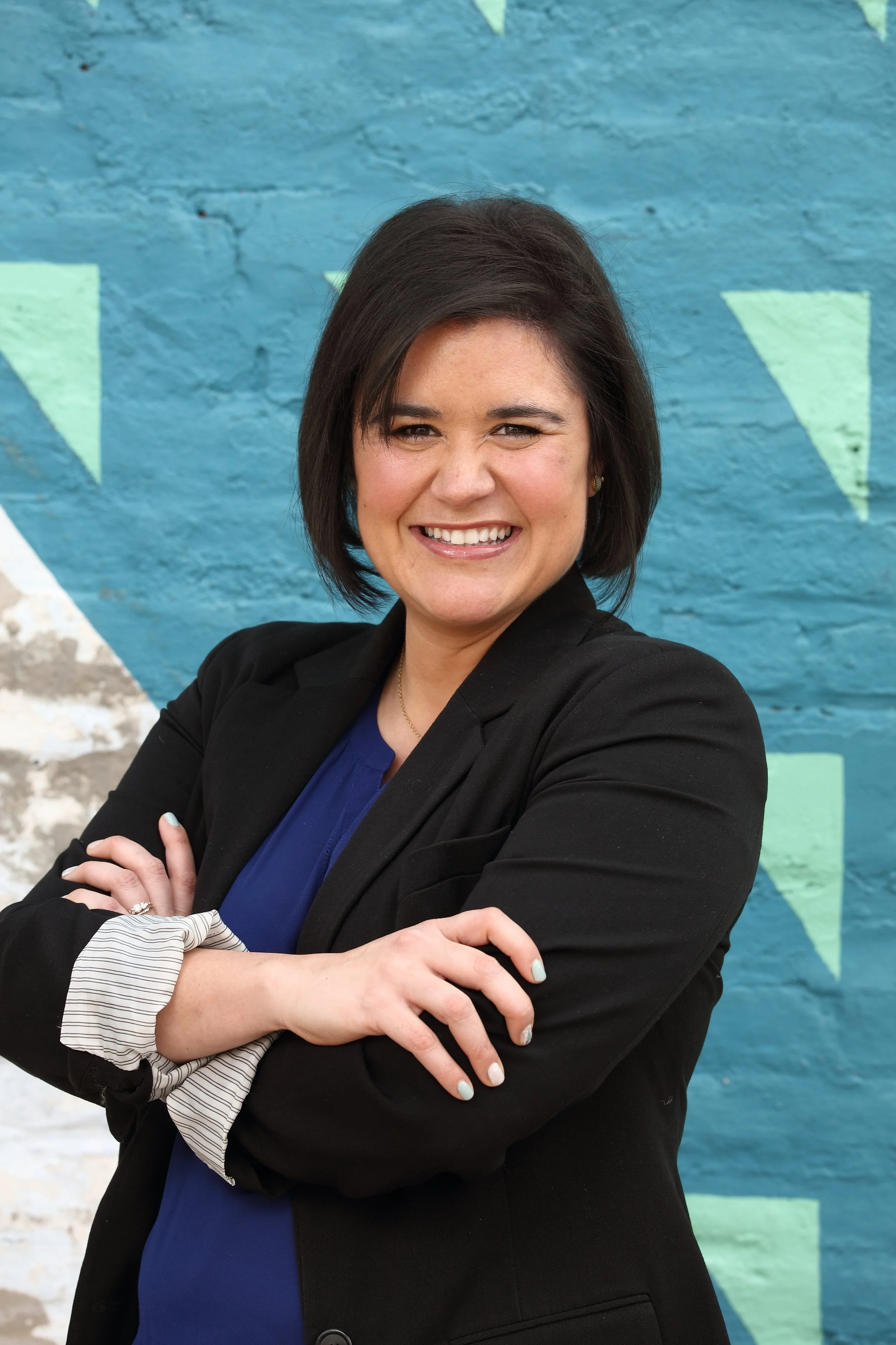 A femme brunette with brown eyes smiles joyfully and excitedly at the camera with her arms crossed. She is wearing a black blazer and stands in front of a colorful painted stone wall with light teal and mint green hues.
