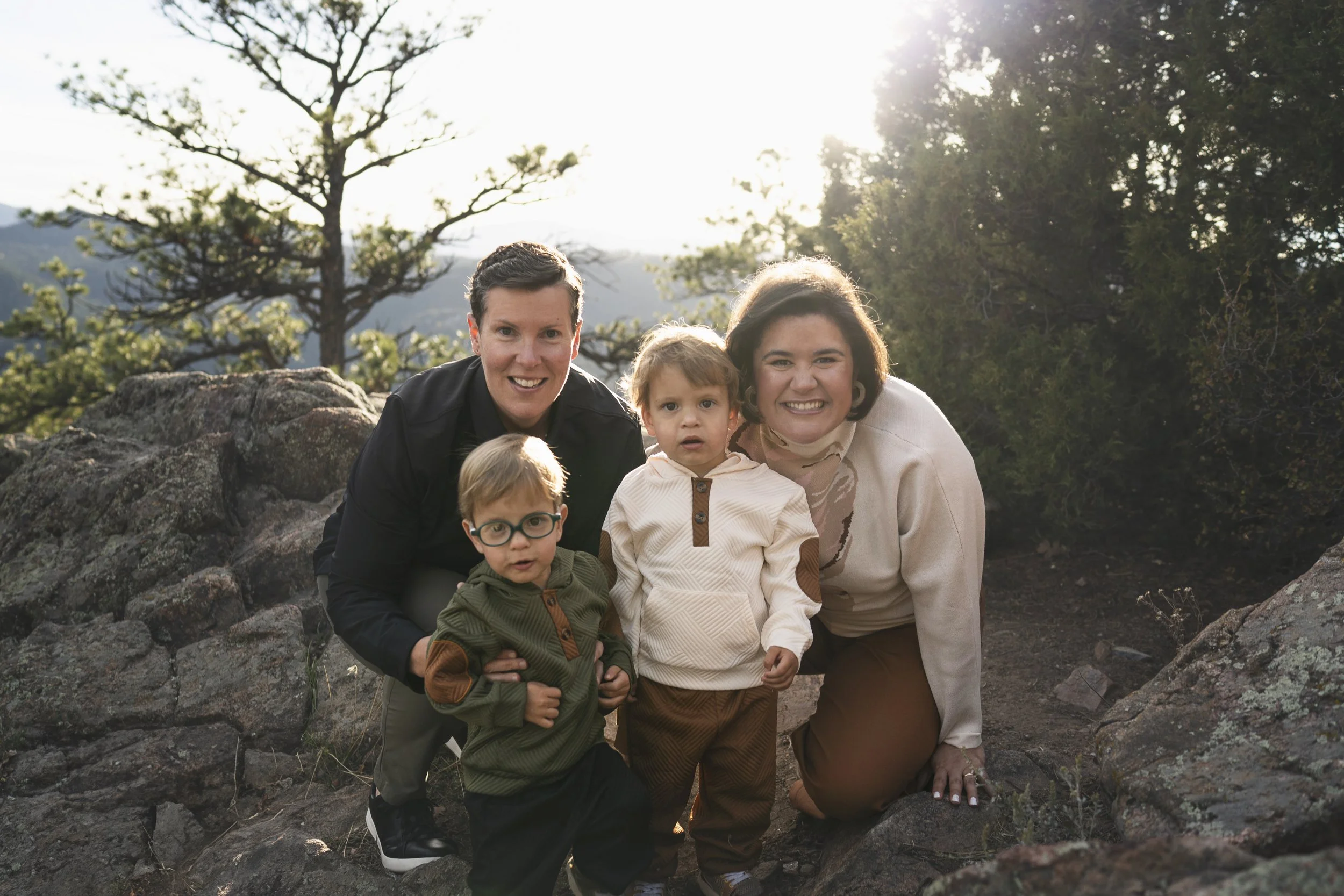 A family of four poses in rocky outcrop at golden hour. Two moms stand closely behind two toddlers.