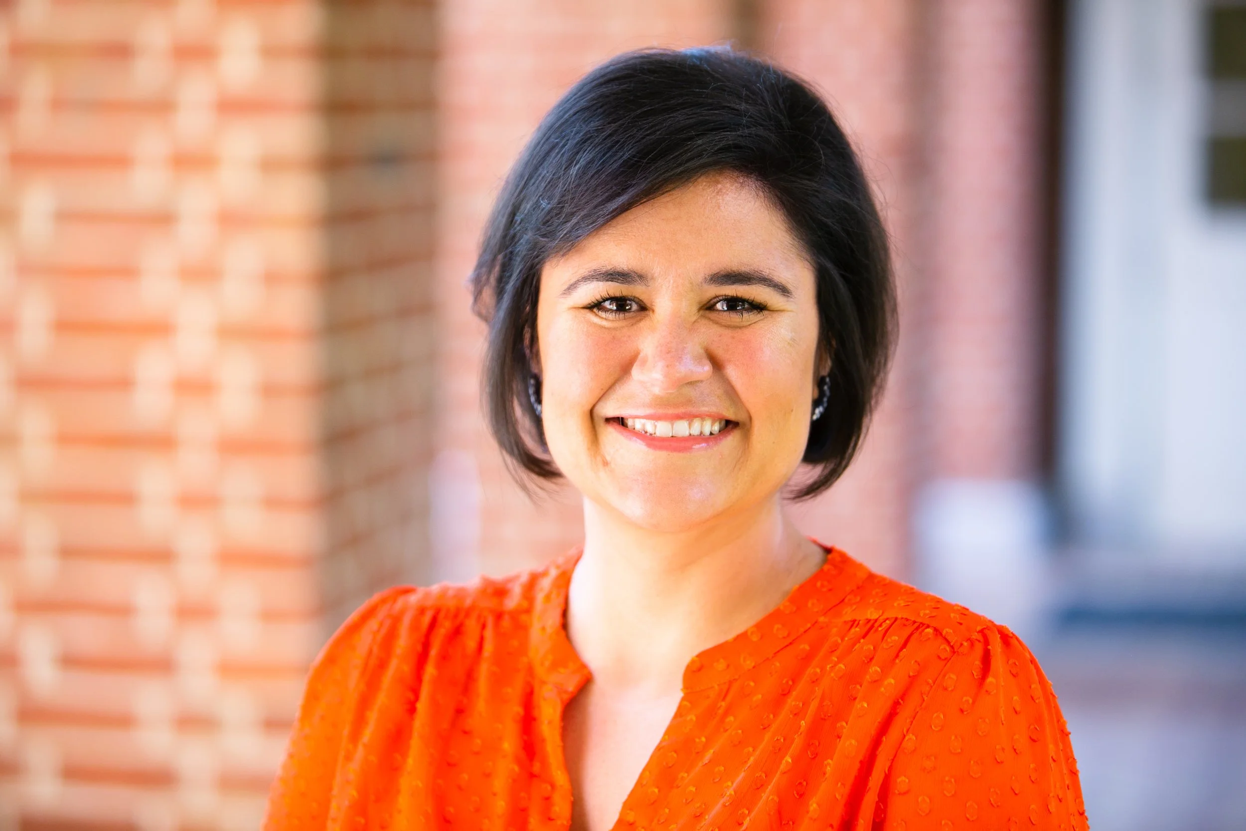 A brunette femme with short layered hair smiles warmly and enthusiastically at the camera. She has on an vivid orange top and stands in front of a red brick wall.